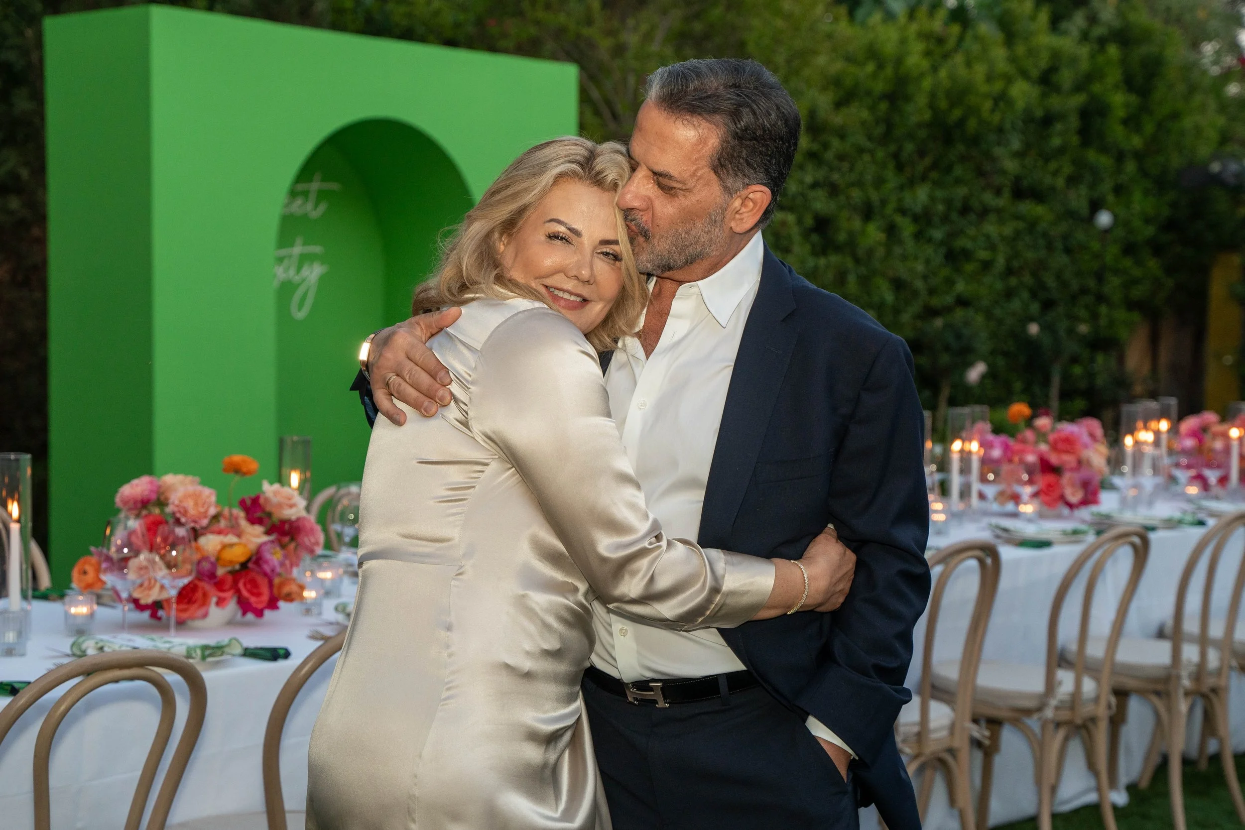 A man and woman hugging at an outdoor event, with tables decorated with flowers and candles in the background.