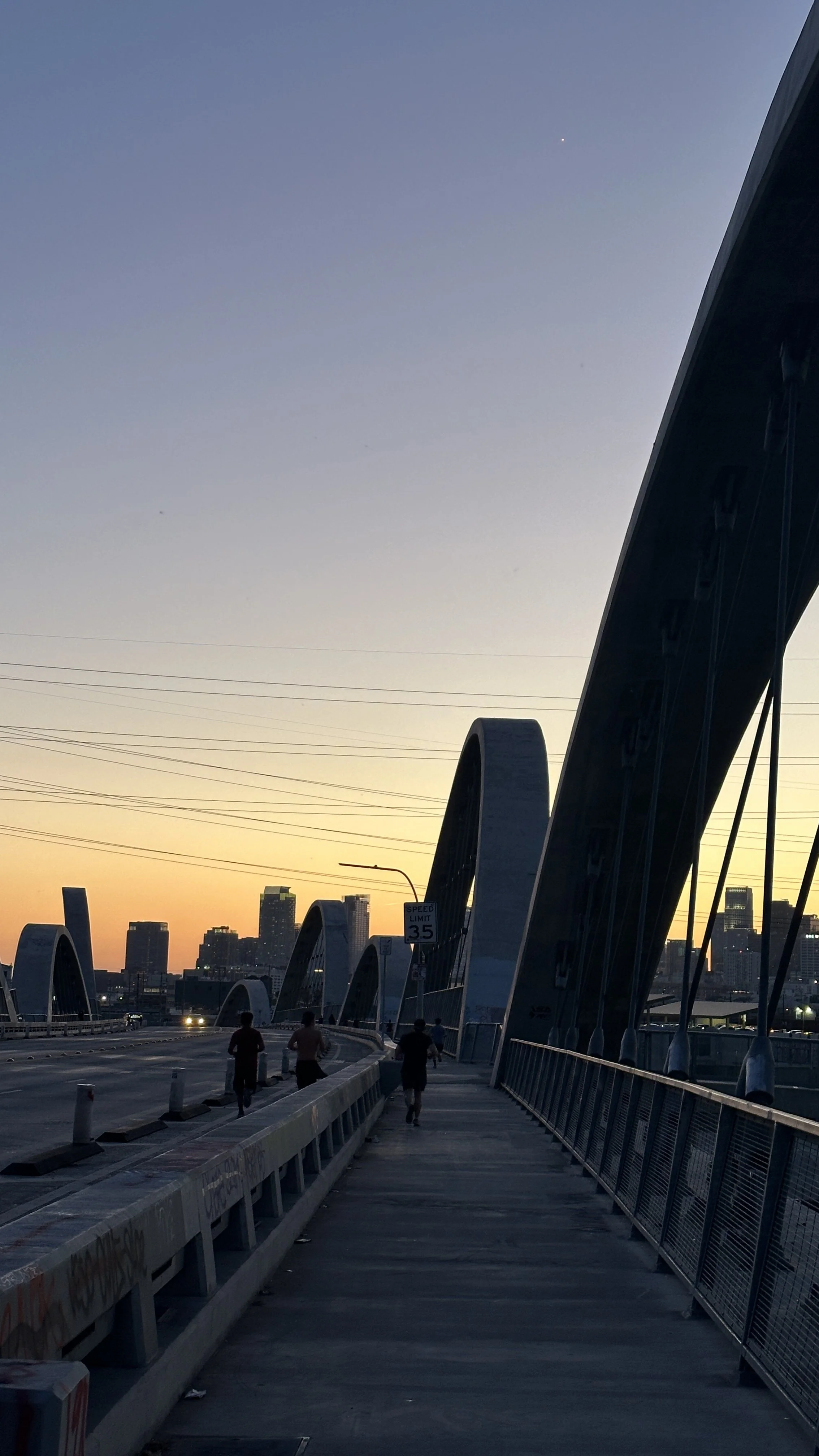 A cityscape at sunset with a bridge in the foreground, showing pedestrians walking and a skyline of tall buildings in the background.