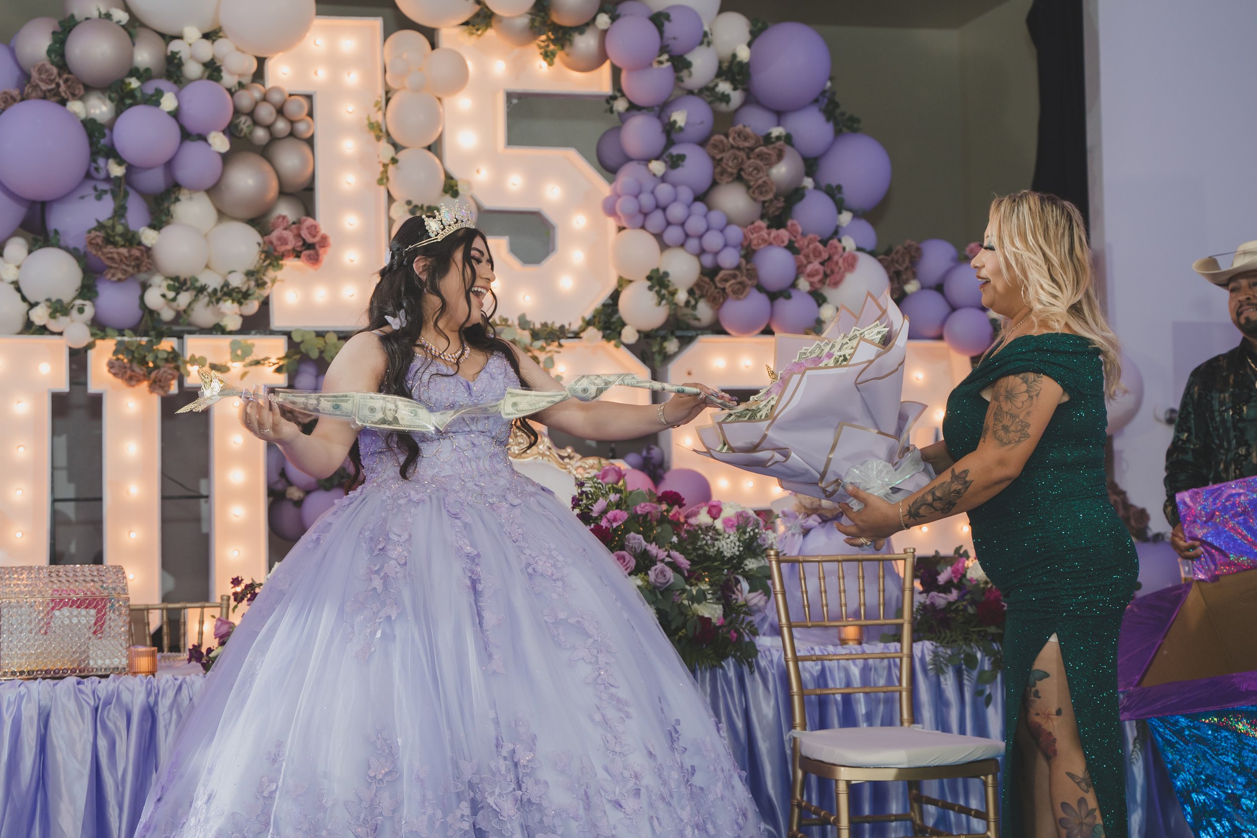 A woman in a lavender ball gown and rhinestone tiara, smiling, is receiving a bouquet of flowers from another woman dressed in a sparkly green dress, in front of a decorated backdrop with purple and white balloons and flowers at a celebration.