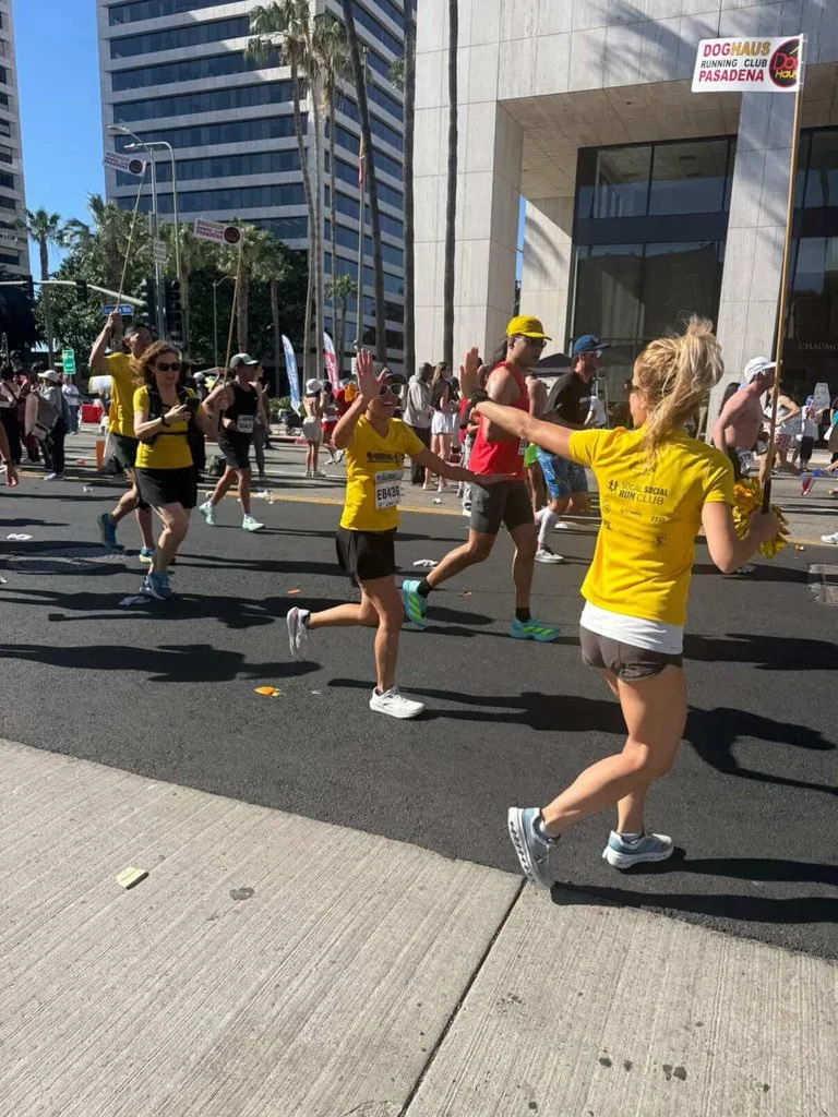 People participating in a street race or marathon, with some giving high-fives, in an urban area with tall buildings and signs reading "Doghaus Running Club Pasadena."