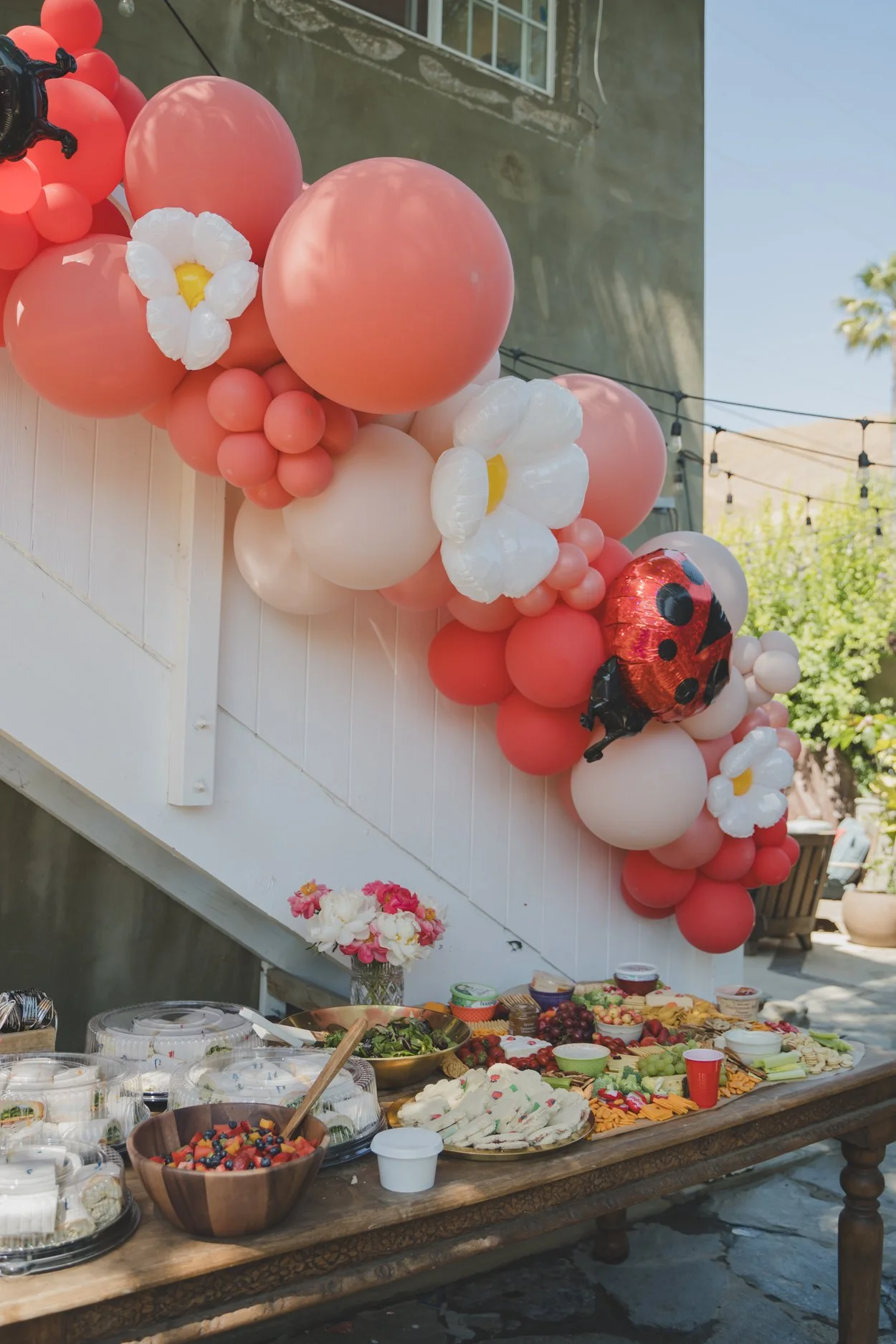 Overflowing table of assorted foods including fruits, cheeses, and salads, with a bouquet of pink and white flowers arranged in a glass vase on the side. Above, a pink and white balloon decoration with ladybug-themed balloons and white flower-shaped 