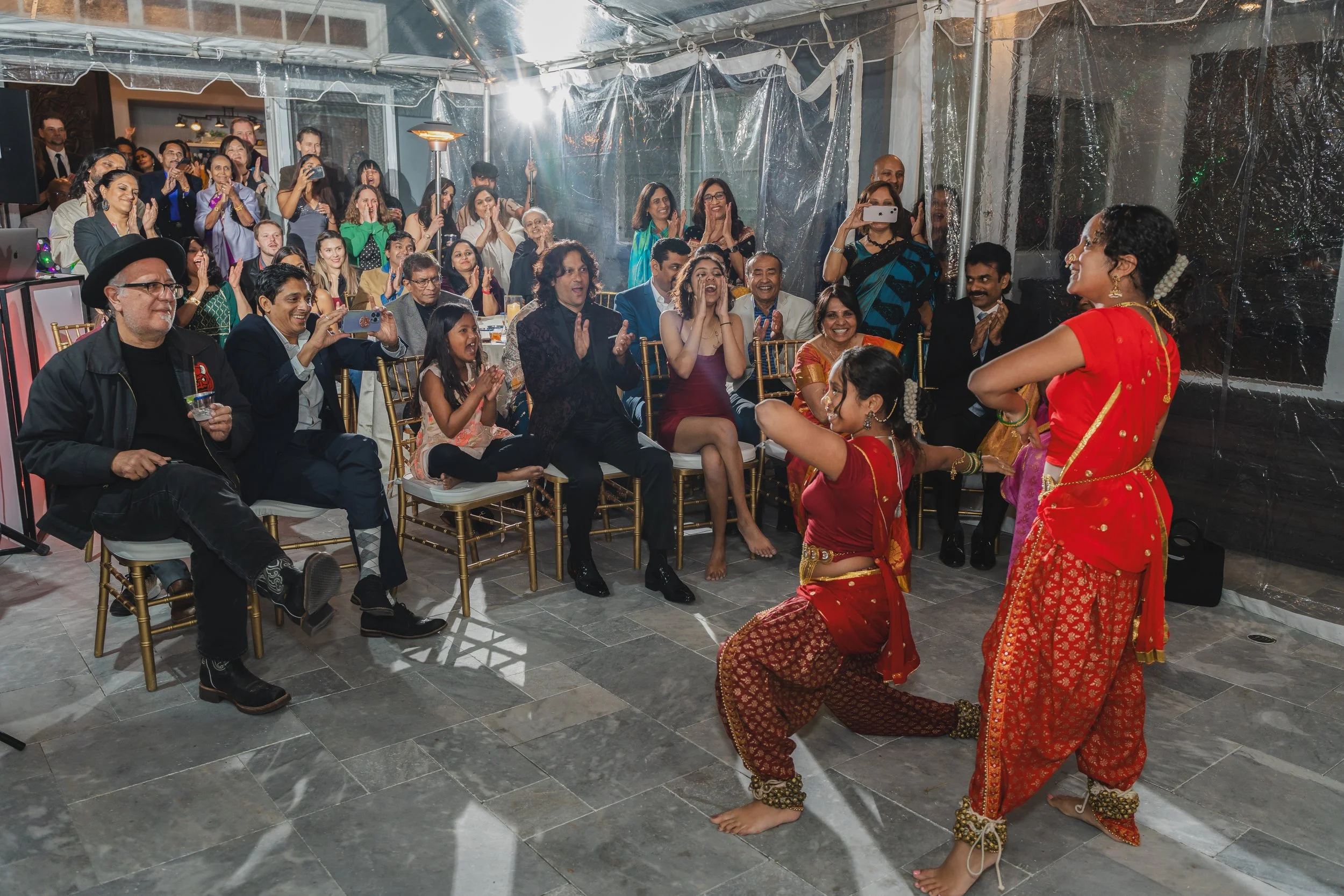 A group of people sitting and standing indoors, watching two women performing traditional Indian dance in bright red and gold costumes. The audience appears engaged and entertained, with some taking photos and videos.