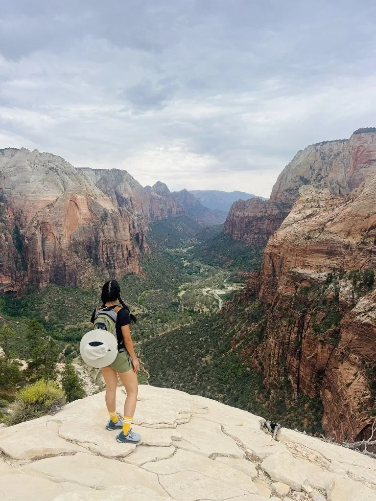 A woman standing on a rocky ledge overlooking a vast canyon with steep cliffs, green vegetation, and a river winding through the valley, under a cloudy sky.