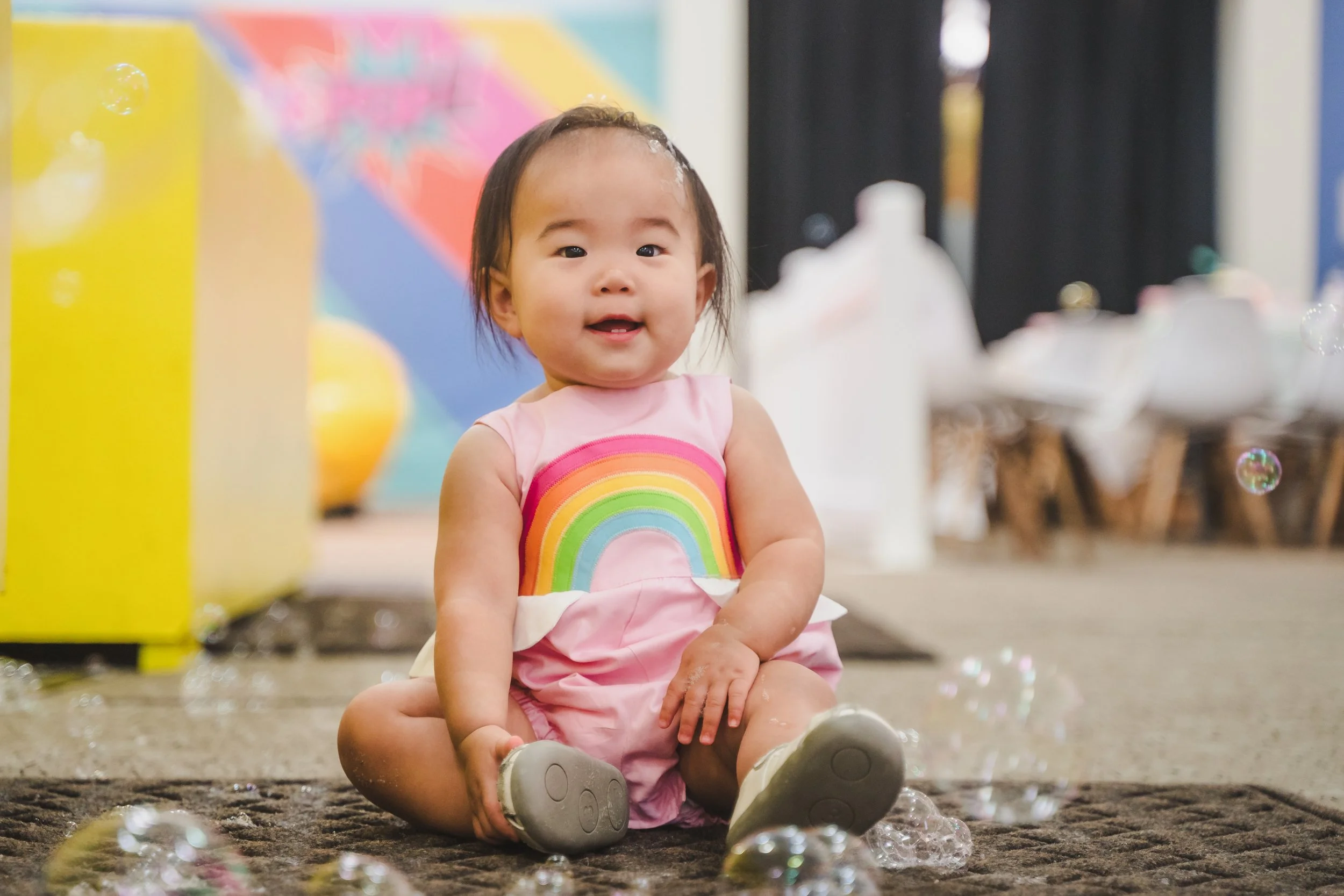A happy young girl sitting on the floor with a rainbow dress, surrounded by soap bubbles, in a room decorated with colorful balloons and streamers.