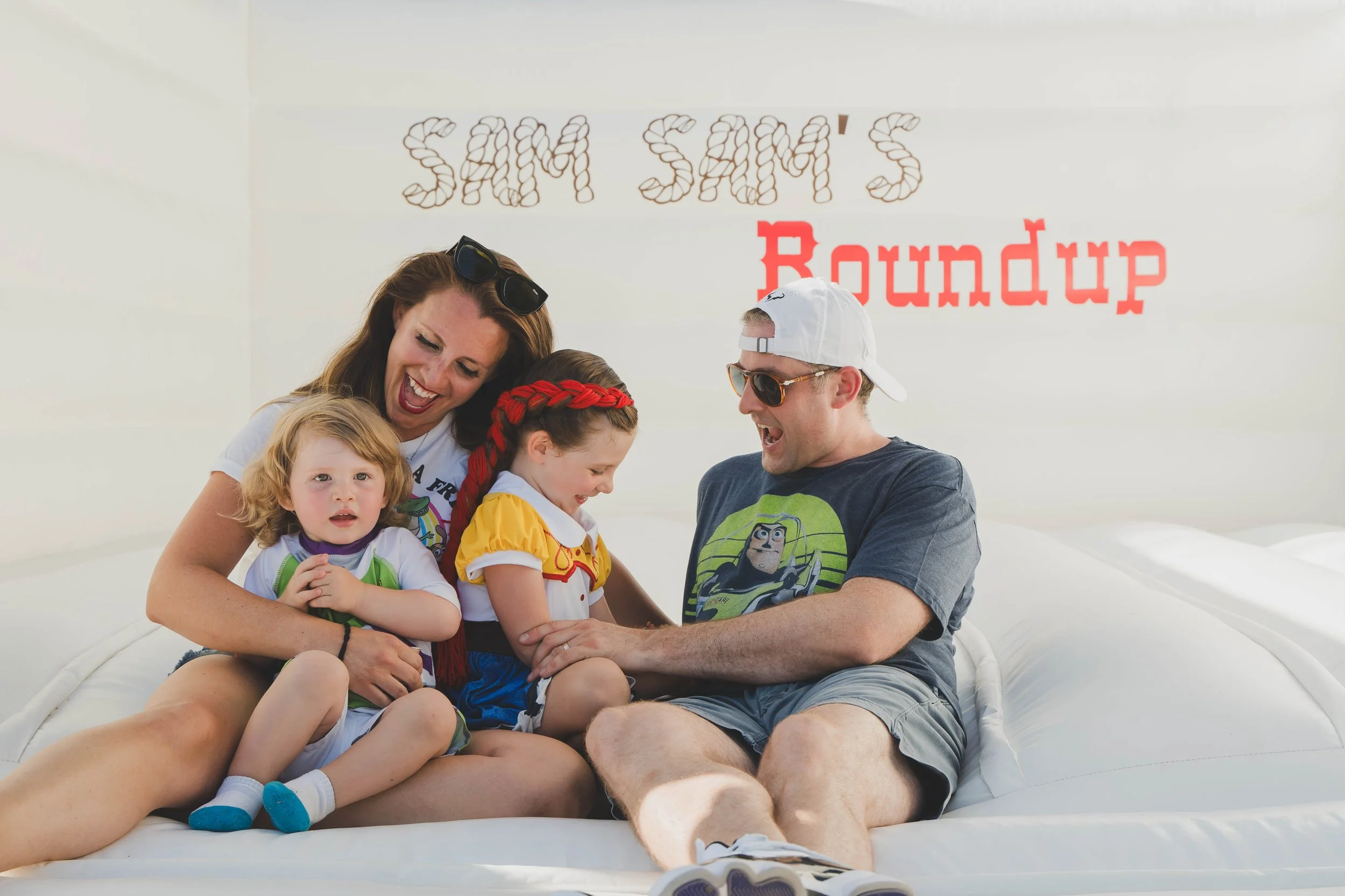 A family of five sitting together on a white inflatable boat, hugging and smiling, with a large white wall in the background that has the words 'SAM'S' in a pattern of coffee beans and 'Roundup' in red text.
