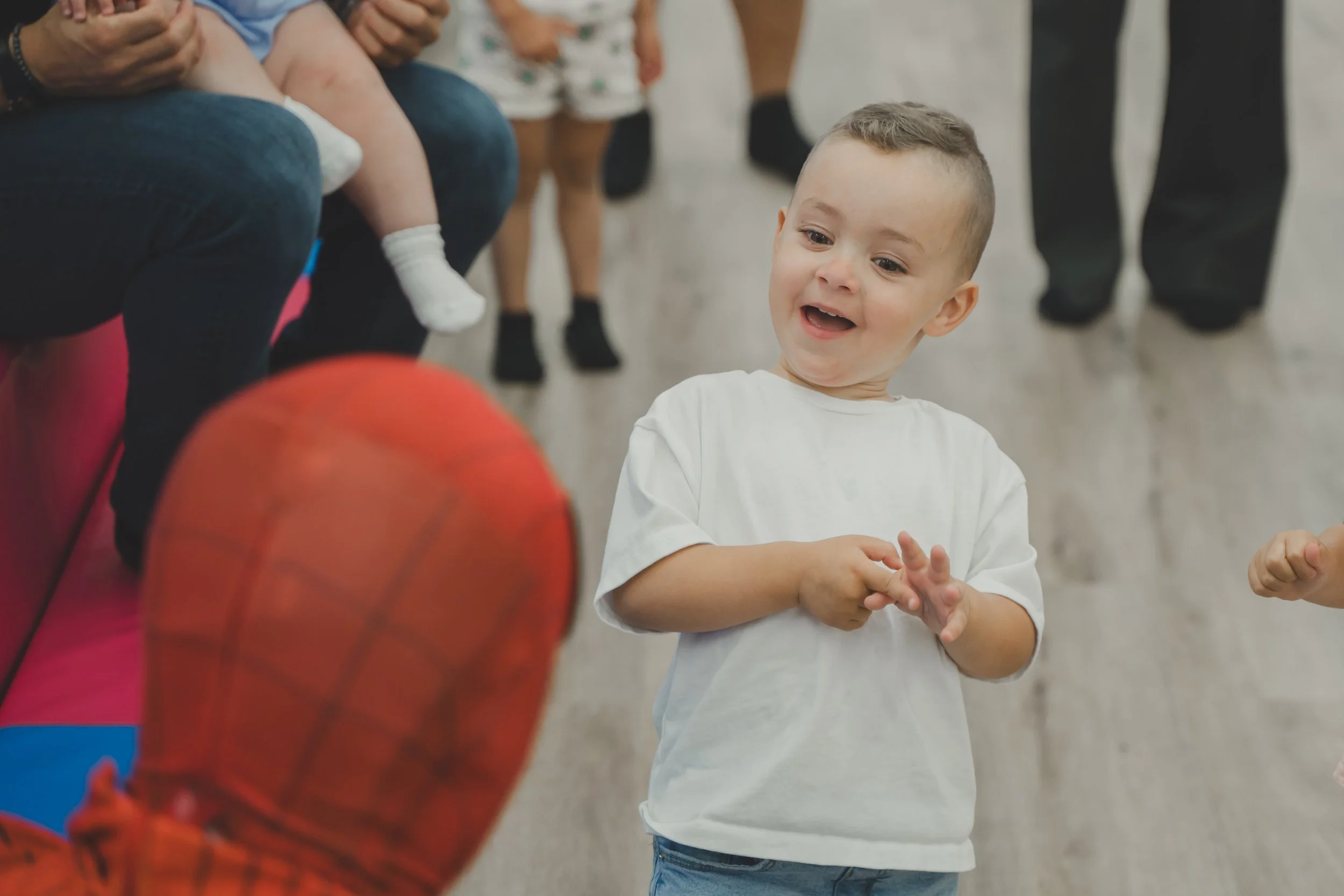 A young boy in a white T-shirt and blue jeans smiling while playing with a red basketball indoors.