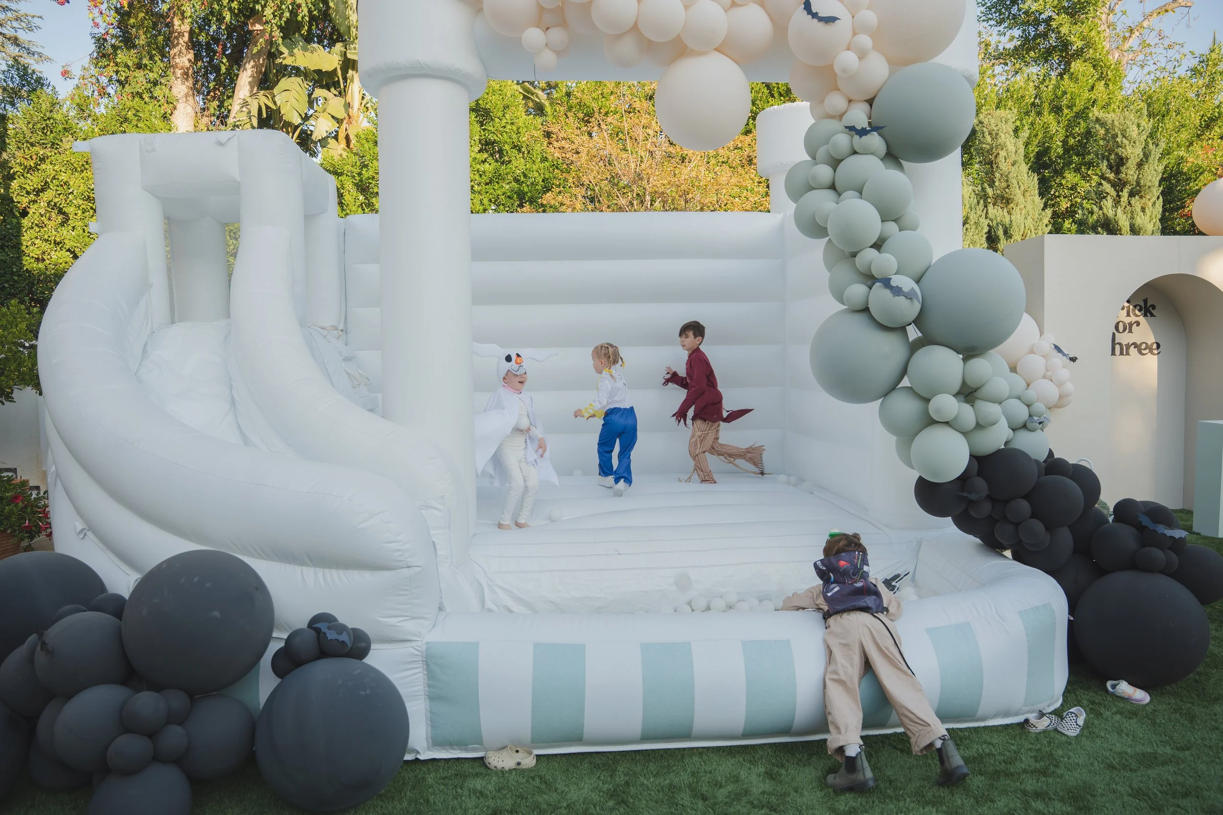 Children playing on a white inflatable bounce house decorated with black, white, and gray balloons and bat cutouts, set outdoors with trees in the background.