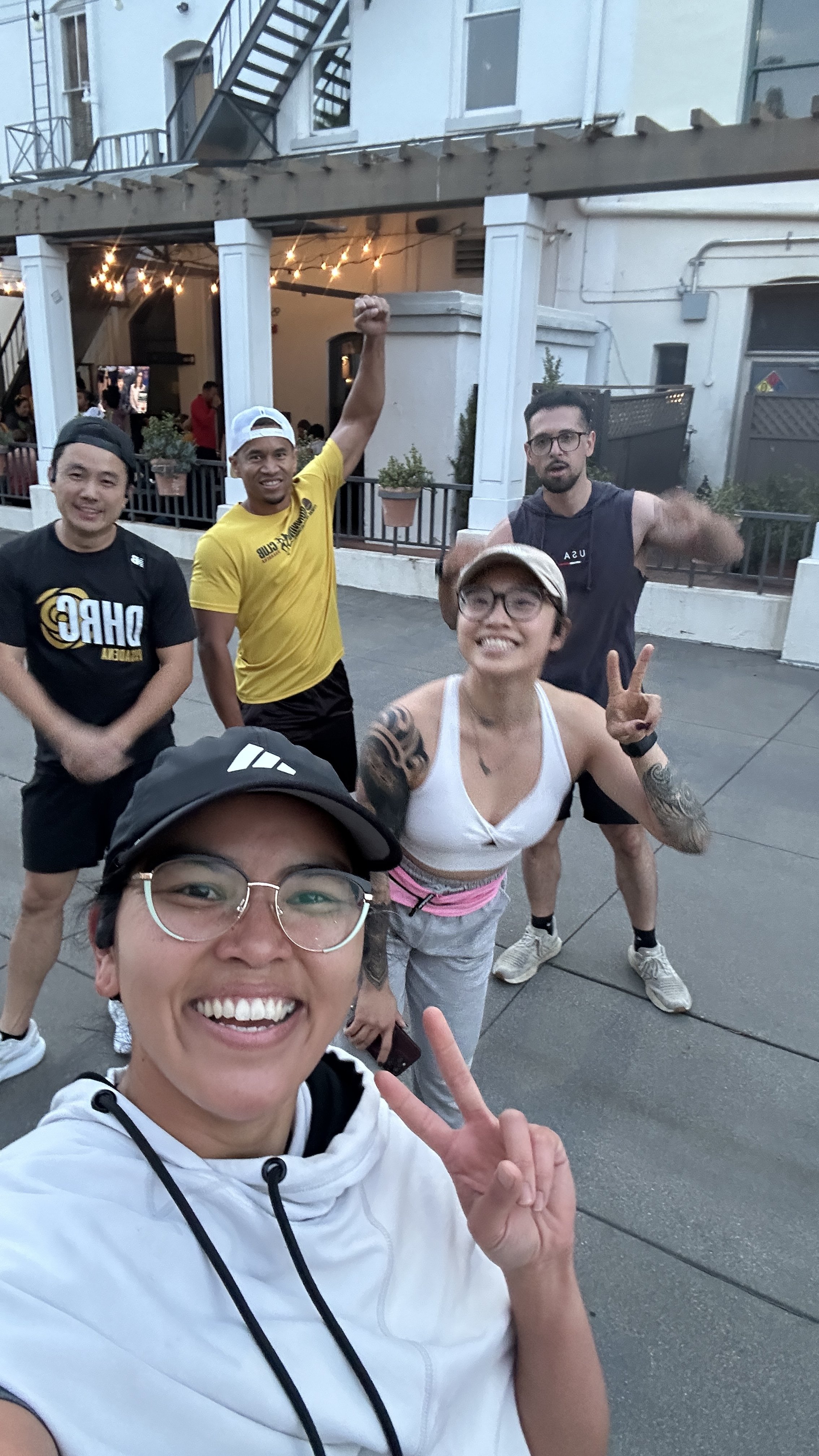 A group of five smiling people, three women and two men, posing for a selfie outside near a building with string lights. The woman in the front is making a peace sign.
