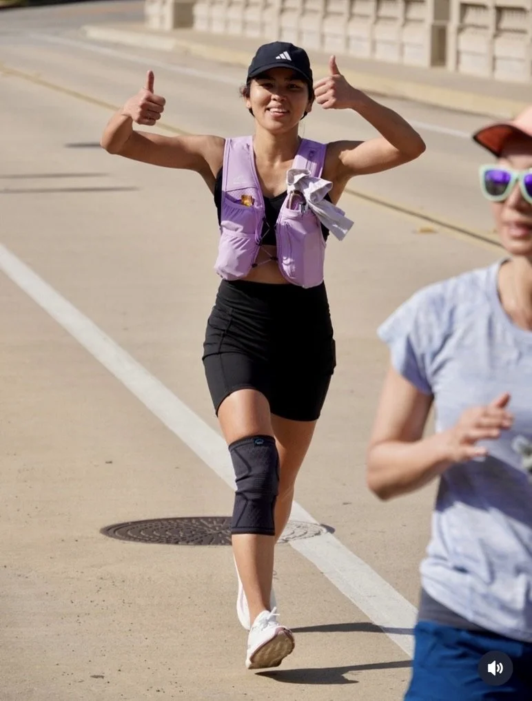Woman wearing a black cap, purple vest, black skirt, and a knee brace, jogging outdoors with a smile and giving thumbs up.