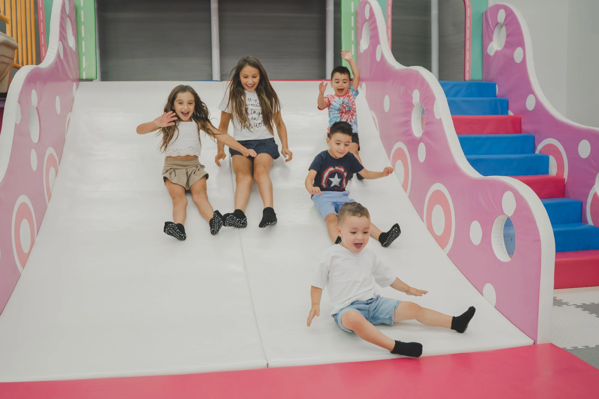 Children sliding down a colorful indoor playground slide with pink and blue steps and pink sides with white polka dots, smiling and having fun.