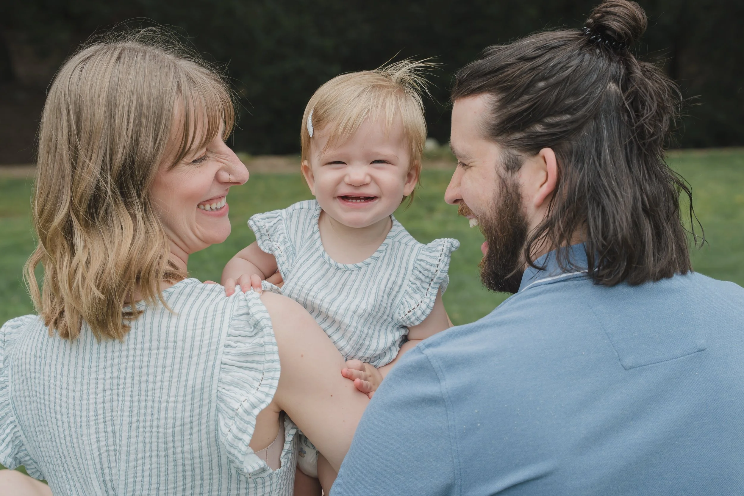 A smiling woman and man hold a laughing toddler outdoors in a park.