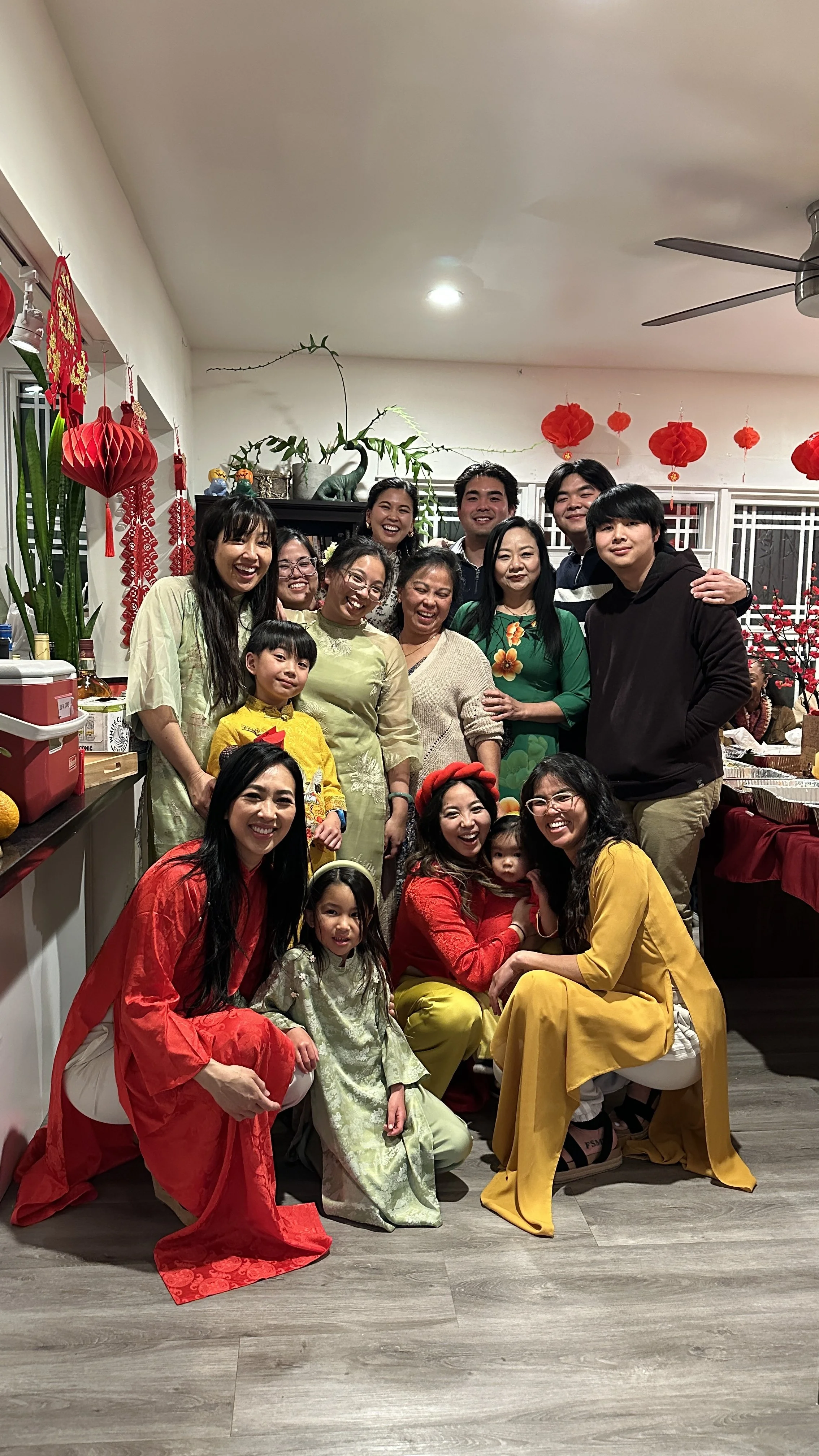 A large group of people, including adults and children, gathered indoors celebrating. Many are dressed in colorful traditional Vietnamese clothing, such as ao dai. The room is decorated with red lanterns, flowers, and festive decorations, indicating 
