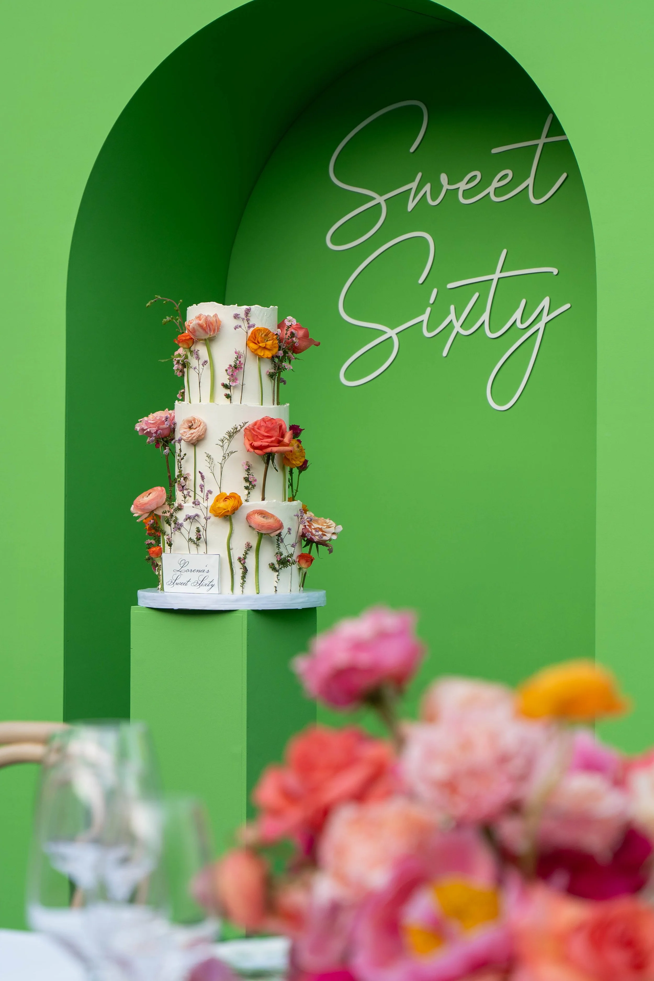 A three-tiered white wedding cake decorated with fresh colorful flowers, placed against a green backdrop with the text 'Sweet Sixty' in a cursive font.
