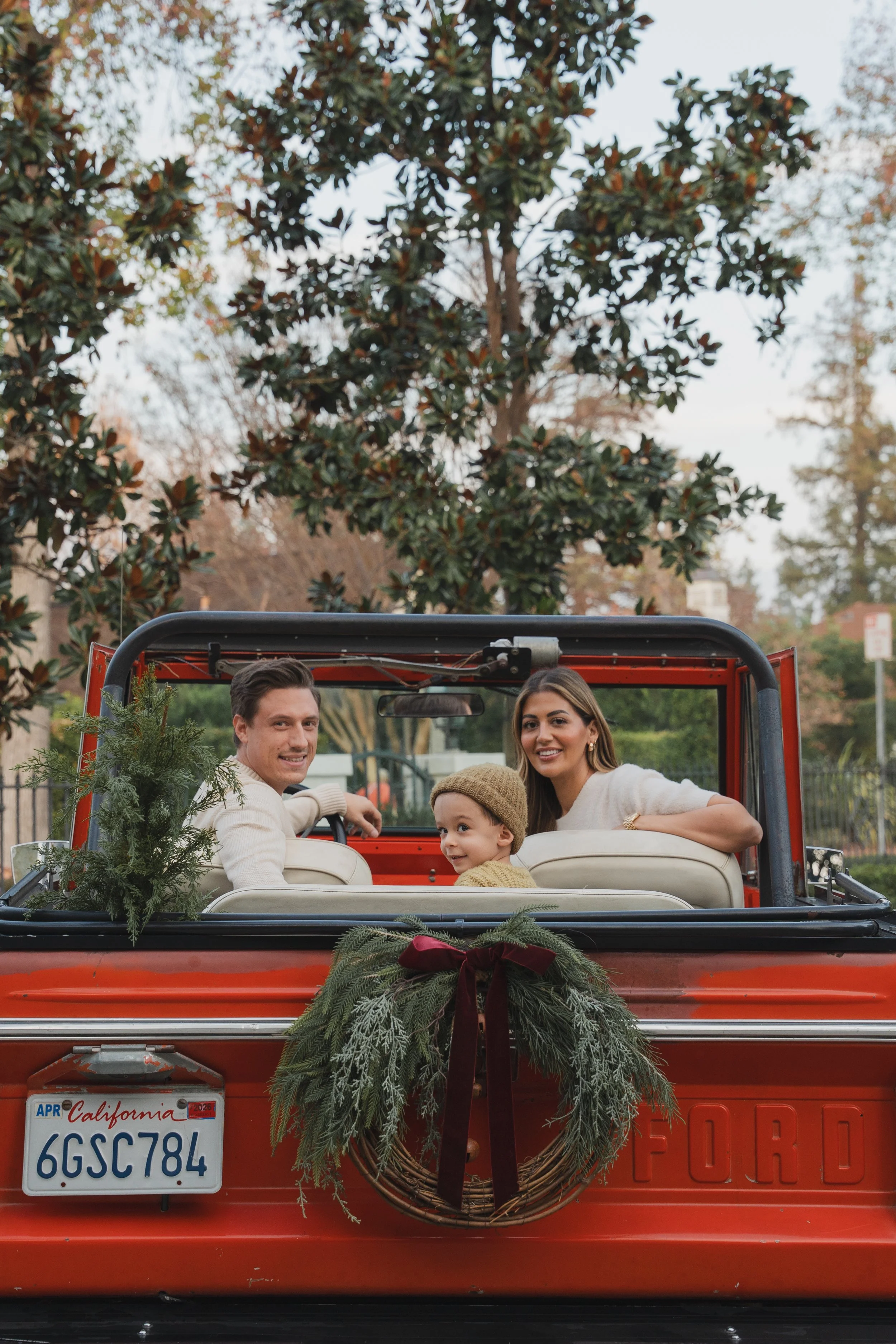 People sitting in the back of a red vintage truck decorated with Christmas greenery and a wreath, outdoors with trees in the background.