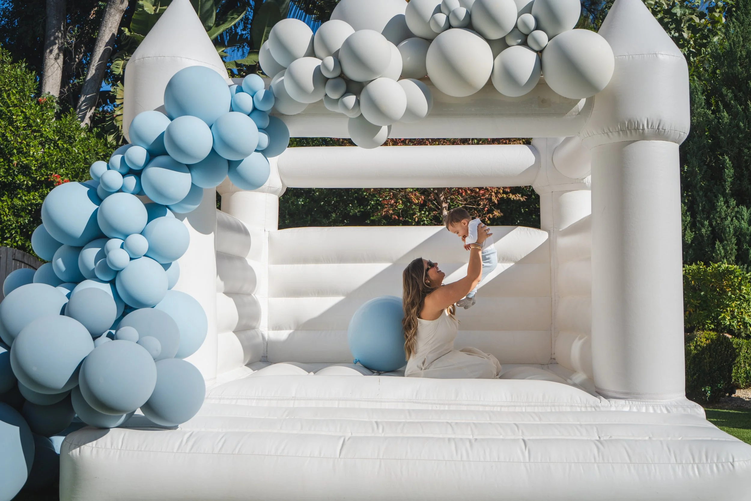 A woman with long brown hair, wearing a white dress, is sitting on an inflatable white structure decorated with light blue and white balloons. She is lifting a young child with brown hair, dressed in white, into the air. The setting appears to be out