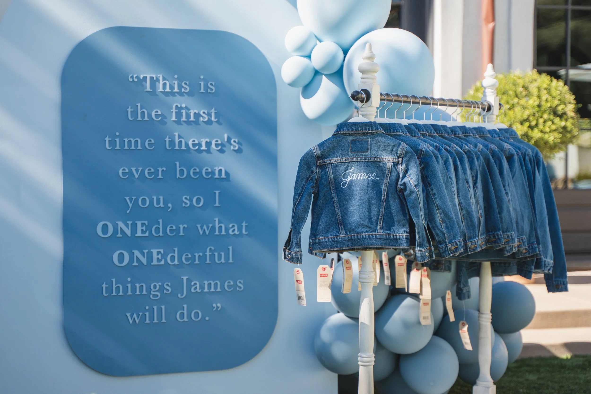 Blue sign with white text quoting James. Next to the sign are blue denim jackets on a rack with white chairs that have balloon-like decorations on top.
