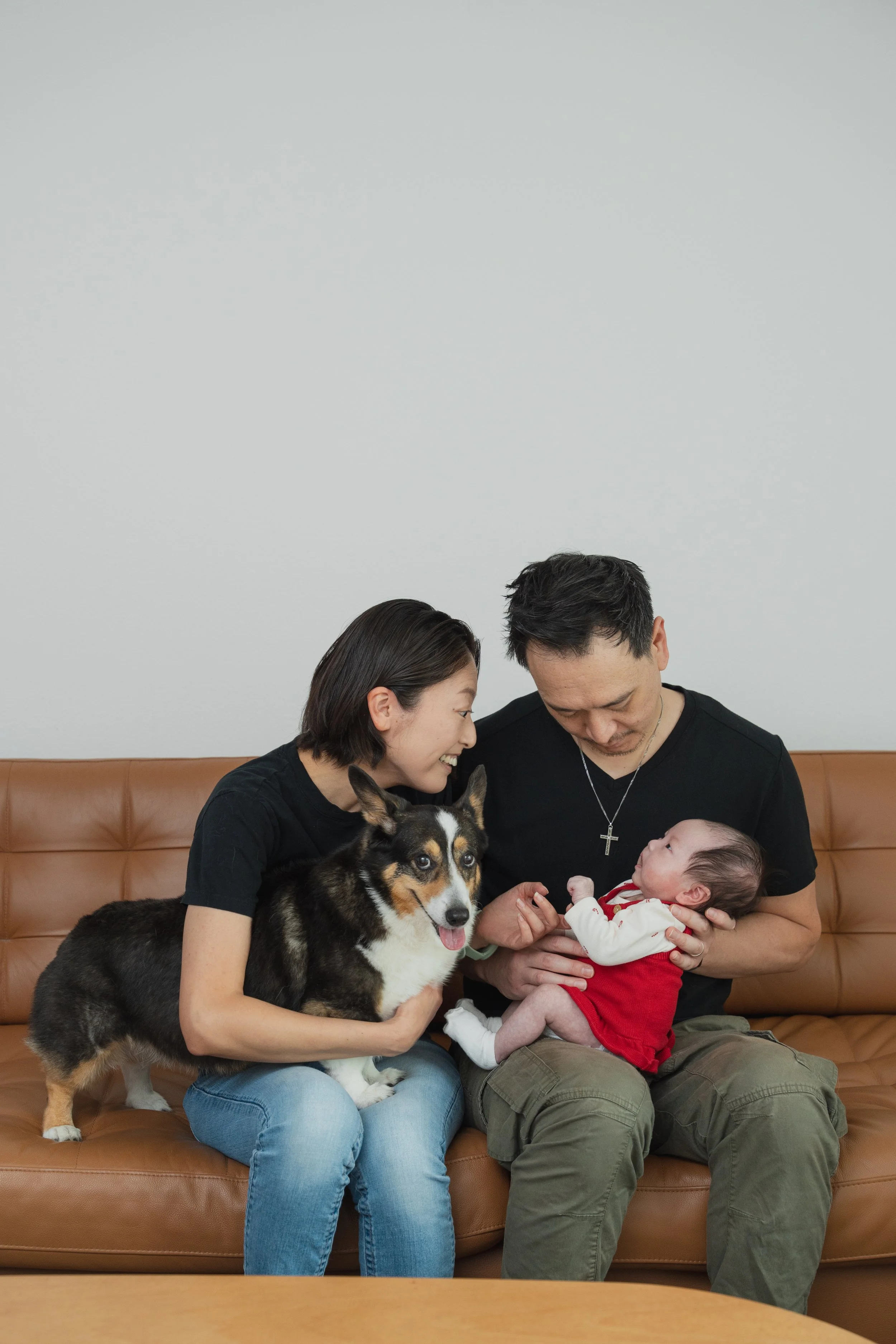 A family sitting on a brown leather couch. The woman is holding a black and white dog, and the man is holding a baby dressed in a red and white outfit. The woman and the man are looking at the baby, who is looking back at them.