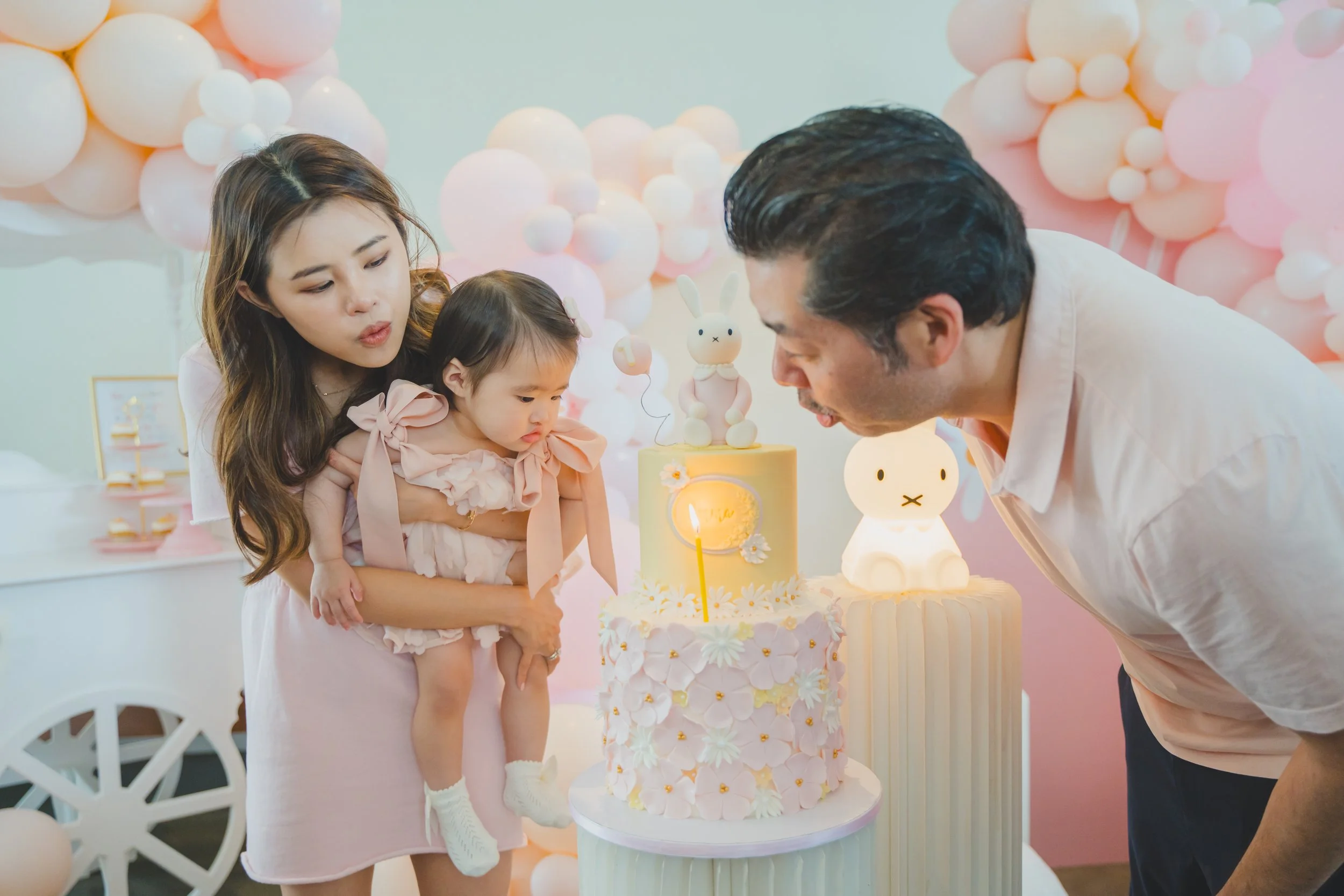 Family celebrating a birthday with a pink and white themed party, cake decorated with bunny-shaped figures, pink and peach balloons in the background, and a young girl in a pink dress.