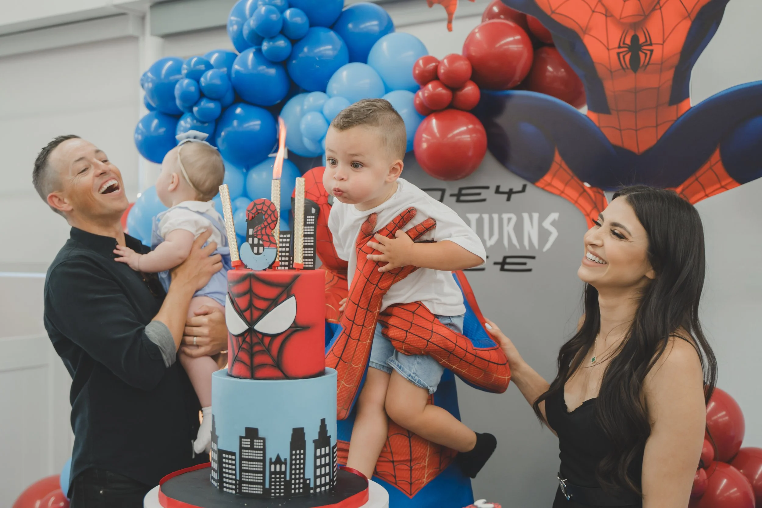 A children’s birthday party with a Spider-Man theme. A boy dressed as Spider-Man is sitting on a woman’s lap, with balloons and a cake decorated with a city skyline and Spider-Man's logo in the background. A man is holding a young girl with balloons 