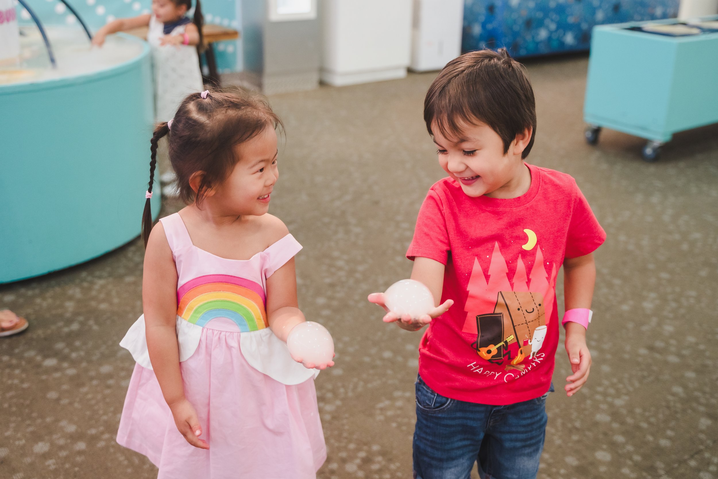 Two young children, a girl with a rainbow dress and a boy with a red shirt, smiling at each other while holding foam balls when celebrating a festive or holiday event.