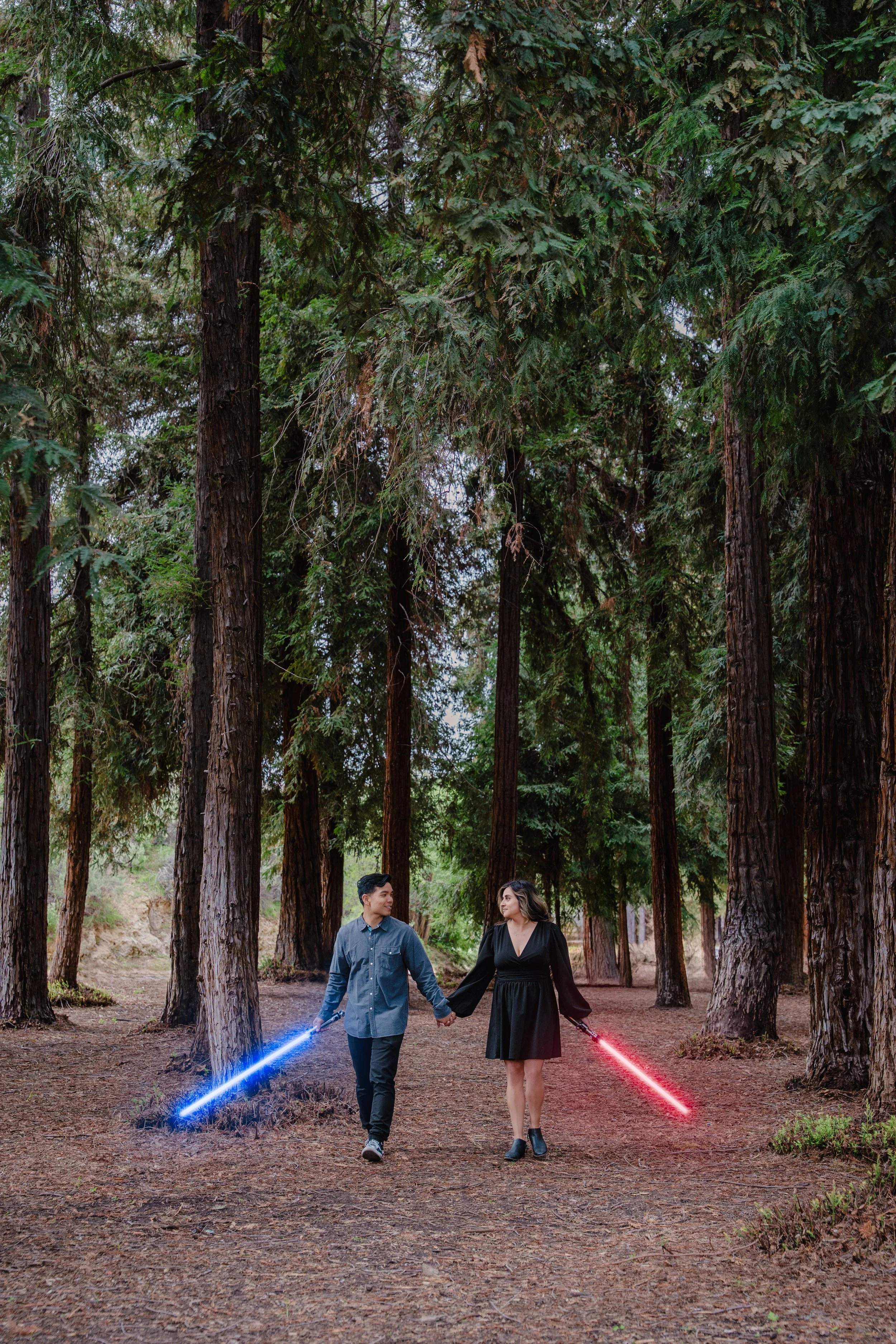 A man and woman holding hands, walking through a wooded forest with tall trees. They are holding toy lightsabers, one glowing blue and the other red, and looking at each other.