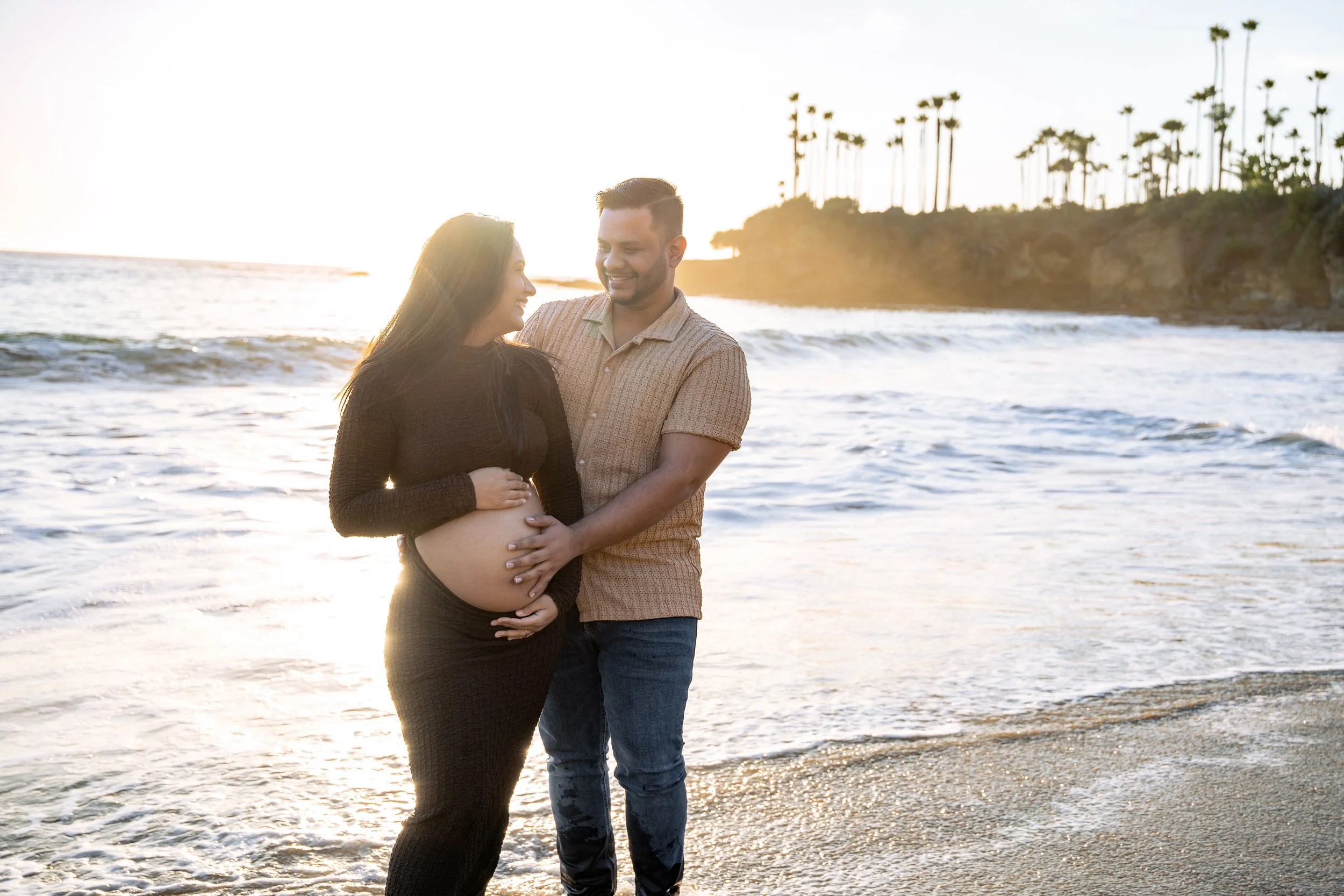 A pregnant woman and a man standing on the beach during sunset, with the woman touching her belly and the man holding her waist, both smiling and looking at each other.