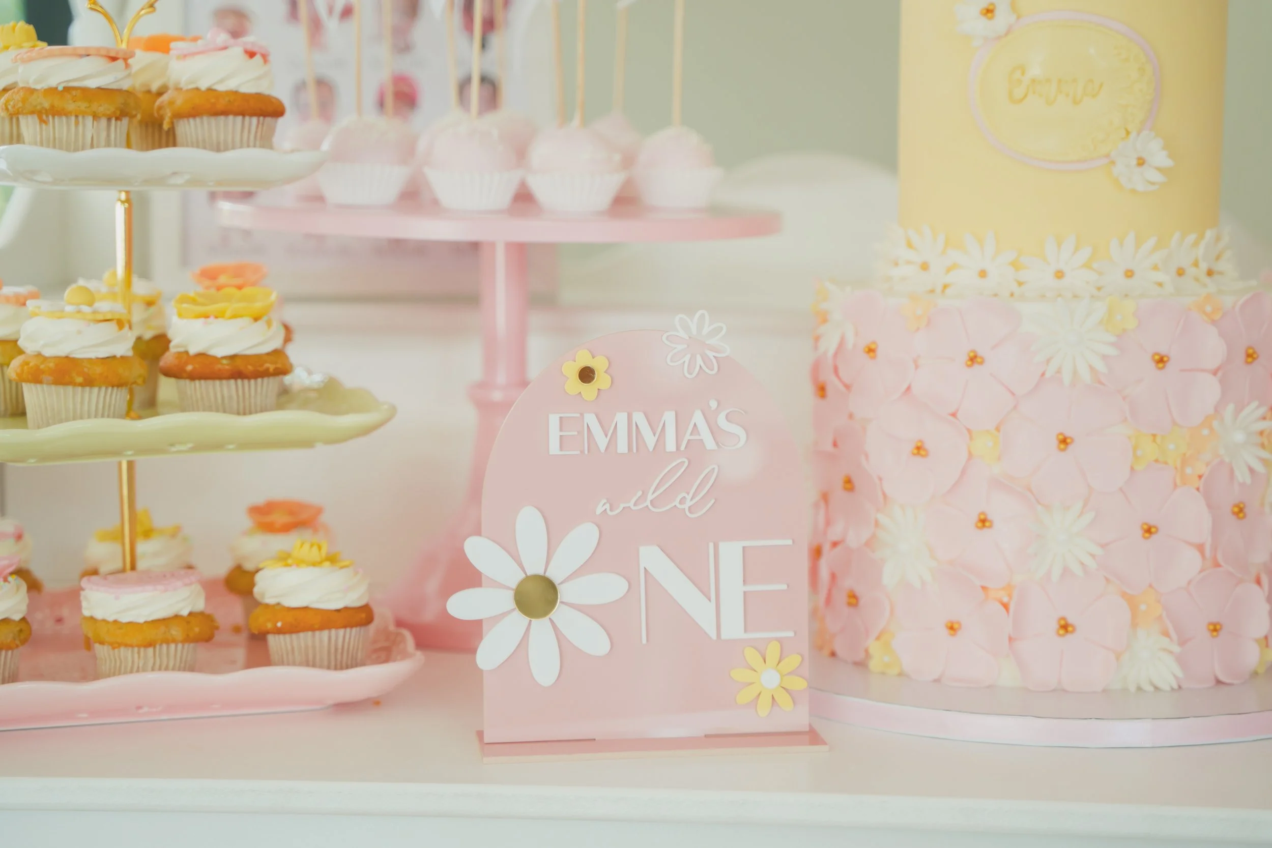 Pastel-colored dessert table with cupcakes, cake, and decorative signs, celebrating a girl's birthday named Emma with a daisy theme.