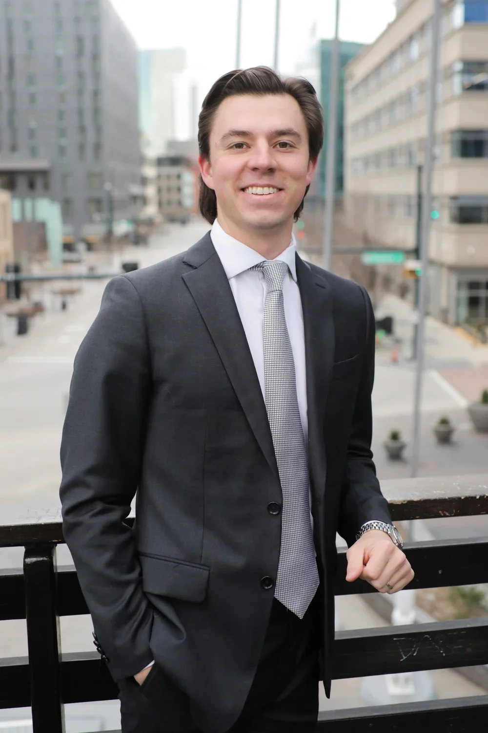 A young man in a dark suit and tie smiling outdoors in an urban setting with tall buildings in the background.
