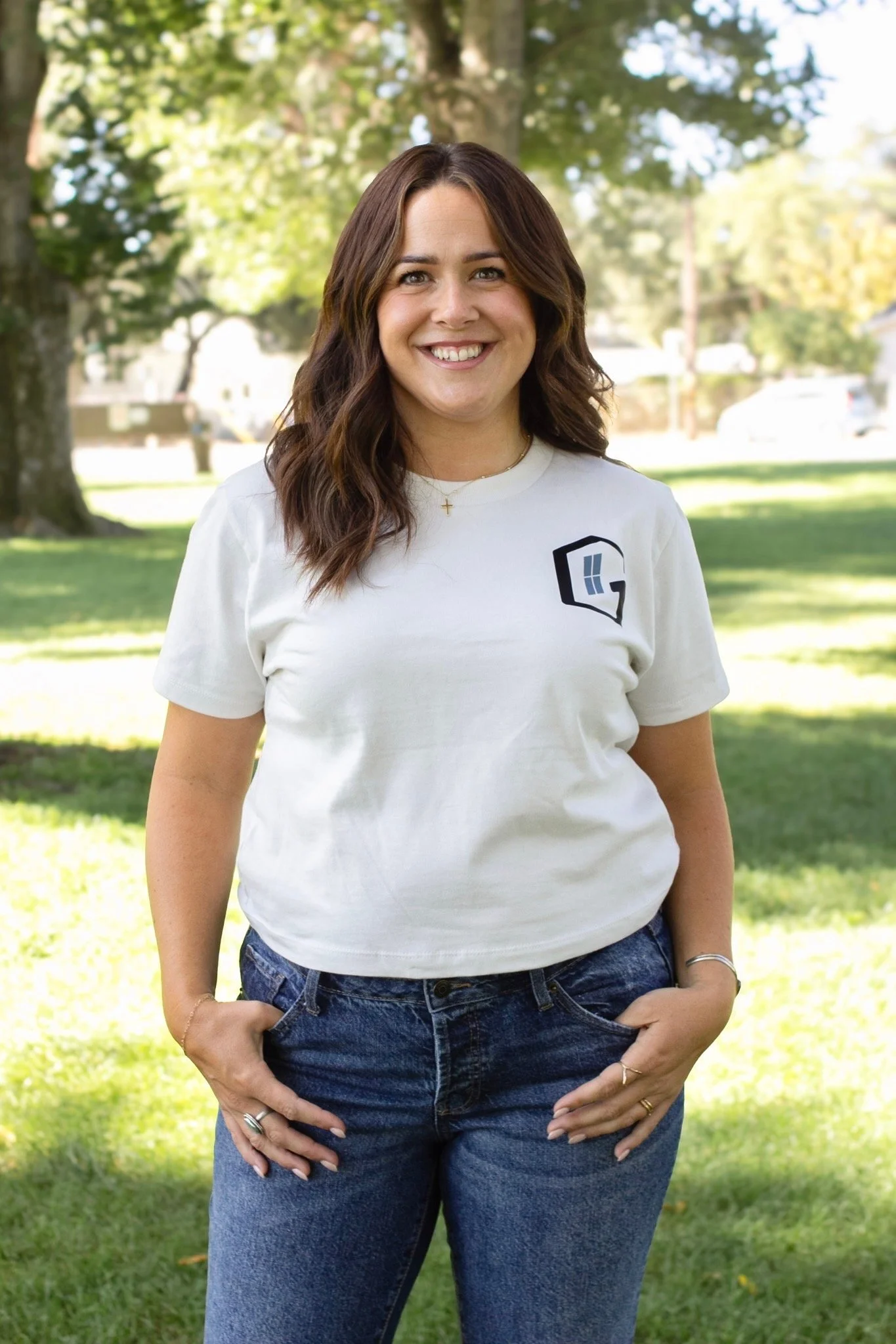 A woman smiling outdoors on a sunny day, wearing a white t-shirt with a logo and blue jeans, standing in a park with trees in the background.