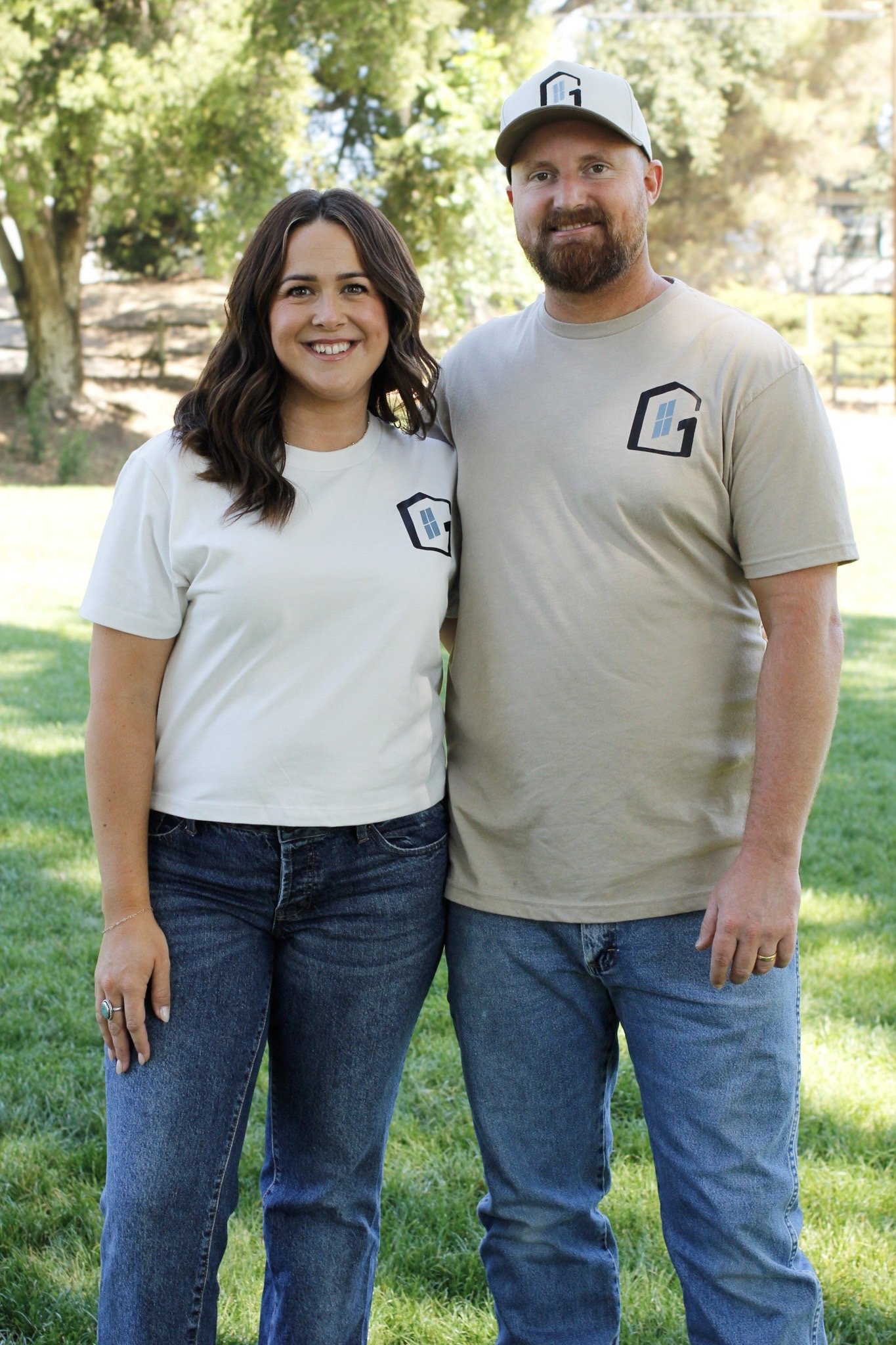 A smiling couple standing outdoors on grass, wearing matching light-colored T-shirts with a house logo, and looking at the camera. The woman has brown hair, and the man has a beard and a cap.