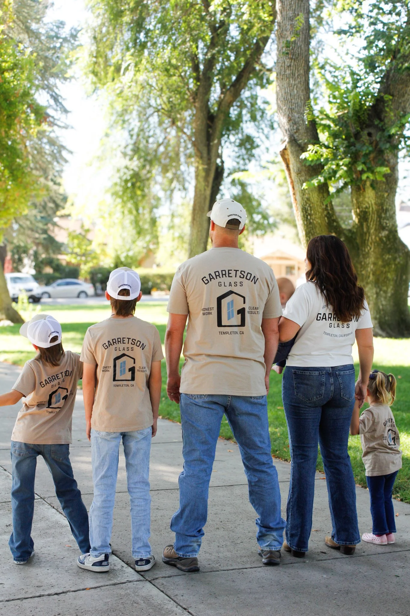 Group of people wearing beige shirts and white caps with 'Garretson Glass' logo, standing outdoors under large trees on a sunny day, possibly at a community event or gathering.