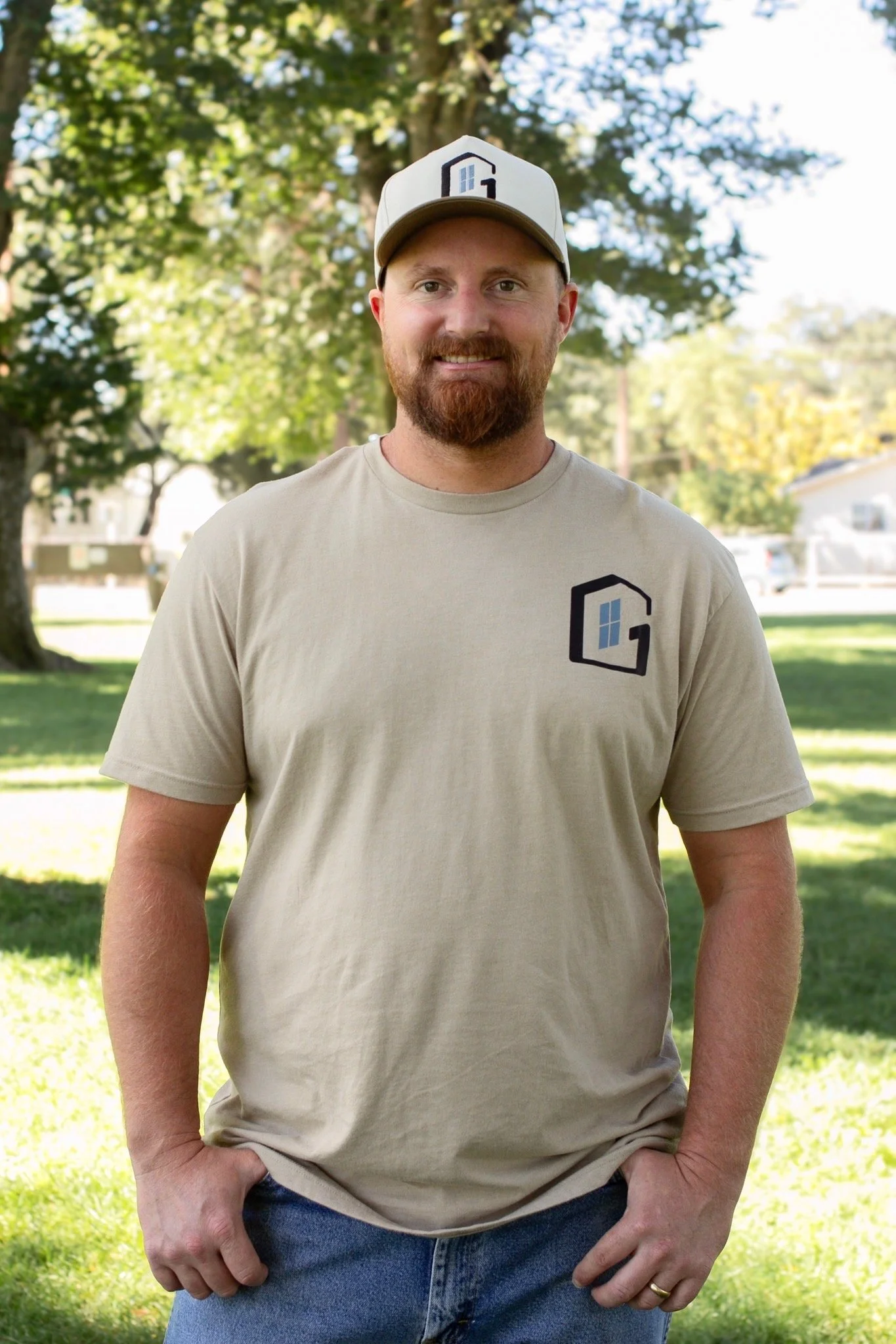 A man with a red beard and mustache wearing a beige t-shirt with a logo and a baseball cap, standing outdoors in a park with trees and houses in the background, on a sunny day.