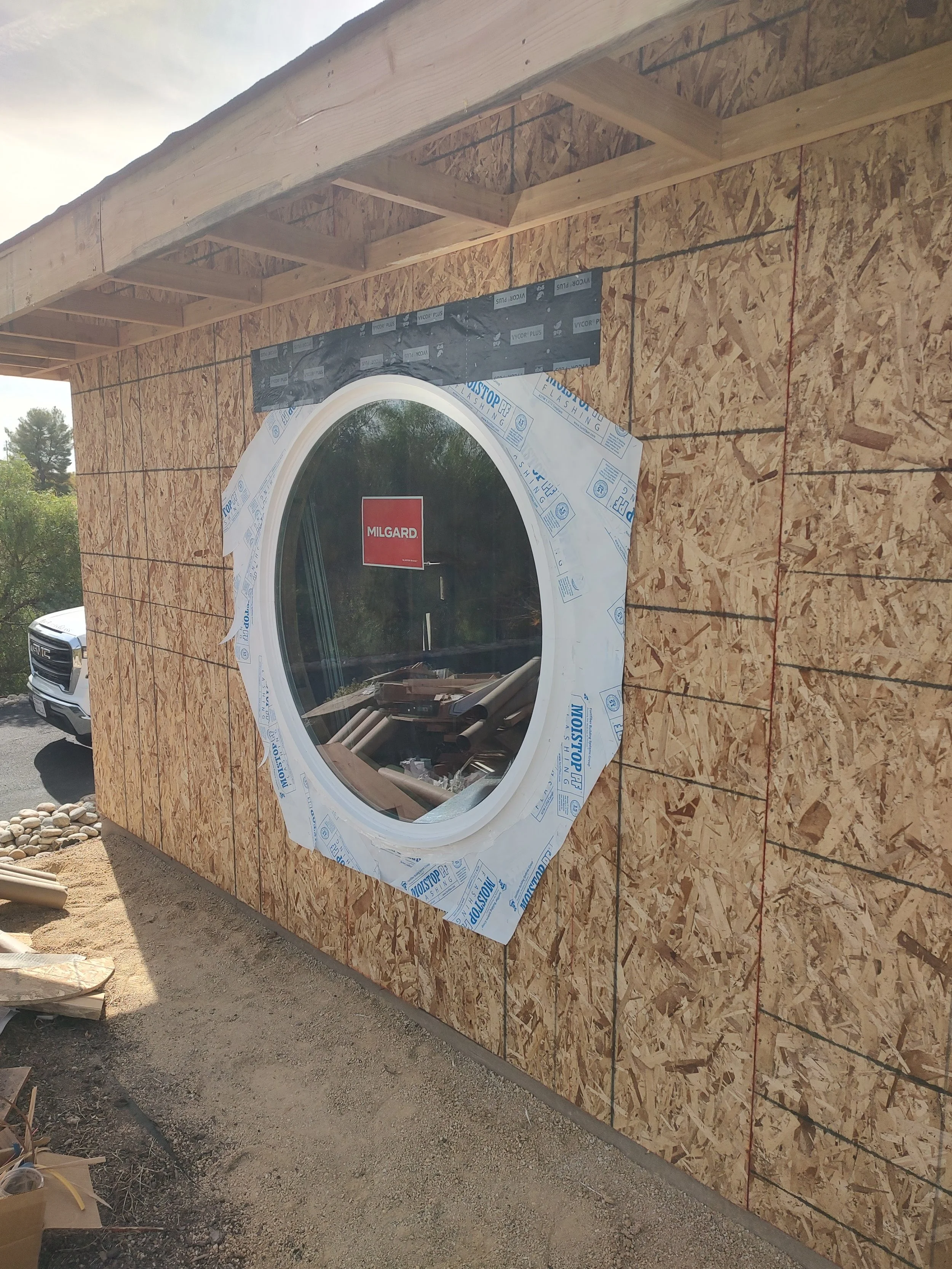 Construction site with a wooden wall with an installed round window, construction materials and a vehicle in the background.