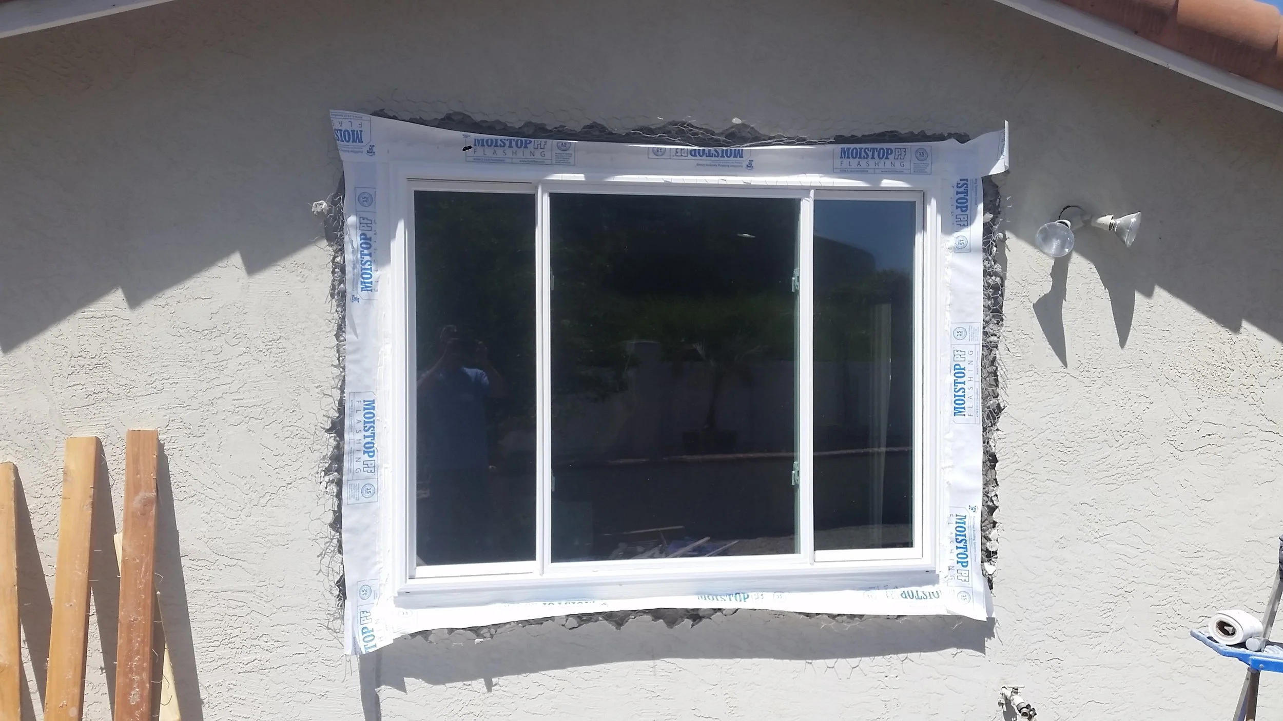 Exterior wall of a house under construction with a window installed, surrounded by white flashing material labeled 'Moistop PF Flashing.' Some wooden boards and construction tools are visible nearby.