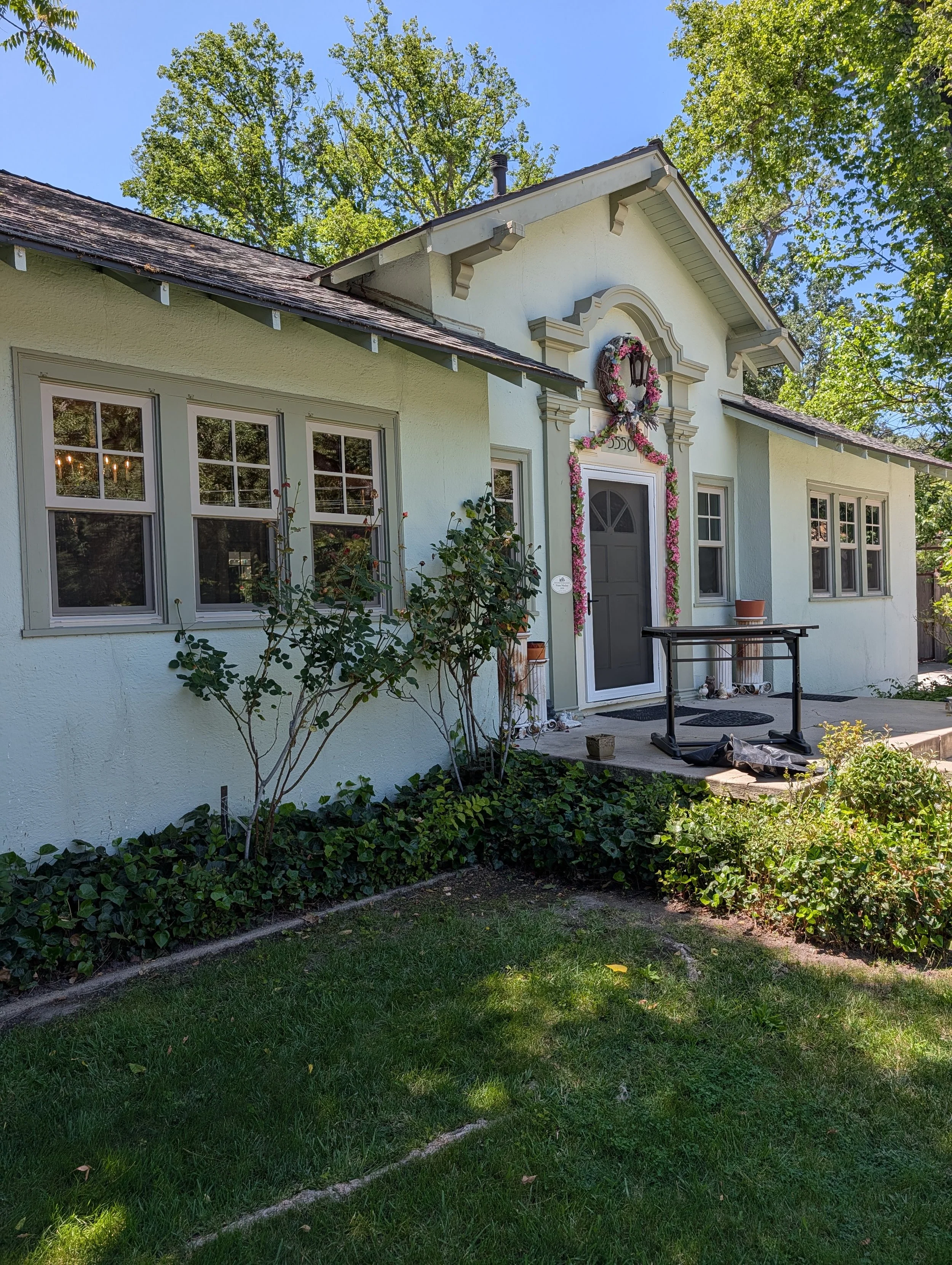 A house decorated for Christmas with pink and white garlands and a wreath, surrounded by green bushes and trees on a sunny day.