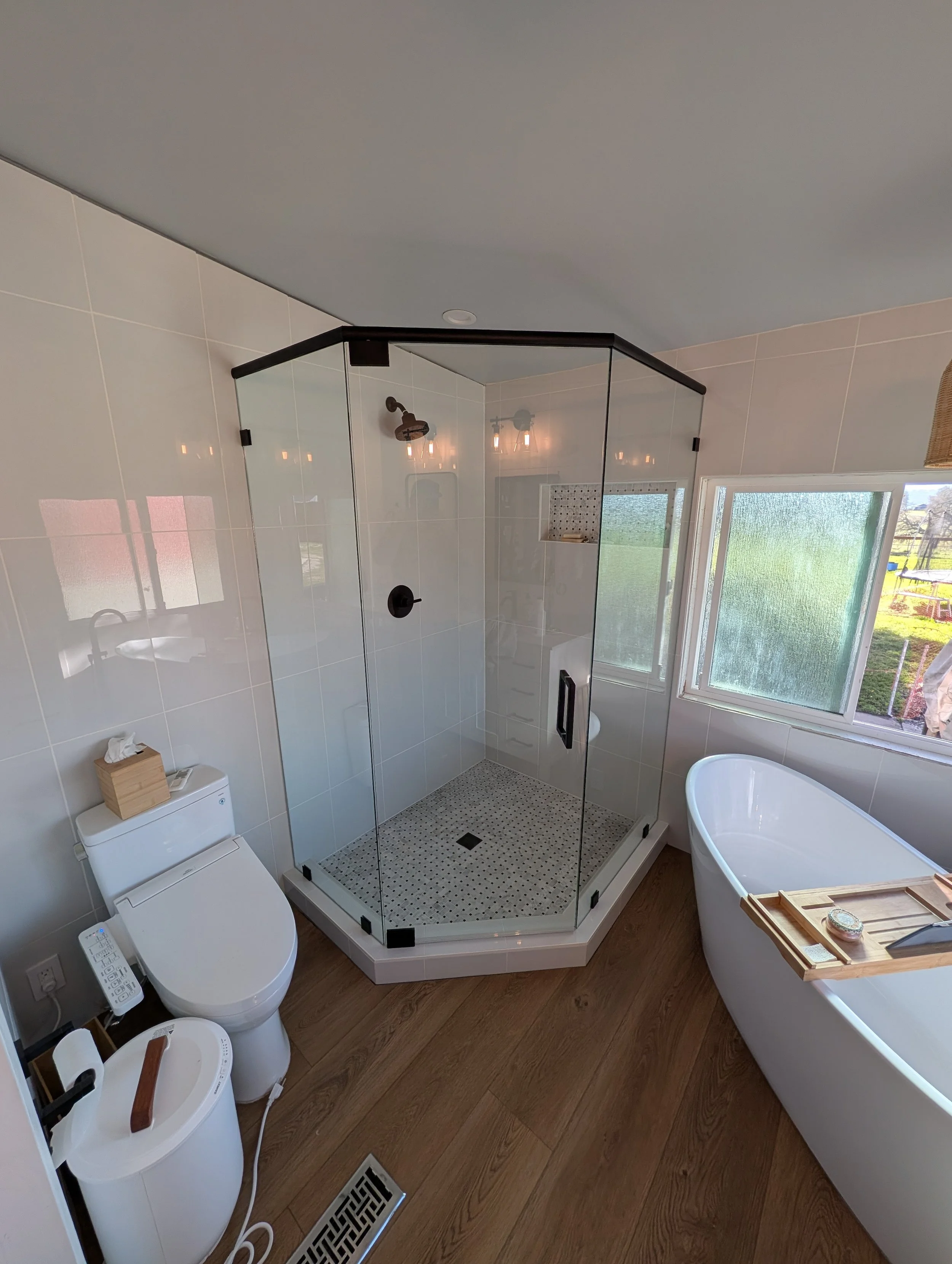 A modern bathroom featuring a glass-enclosed shower with black hardware, a freestanding bathtub with a wooden tray, a wall-mounted toilet with a bidet seat, large windows with natural light, and wooden flooring.