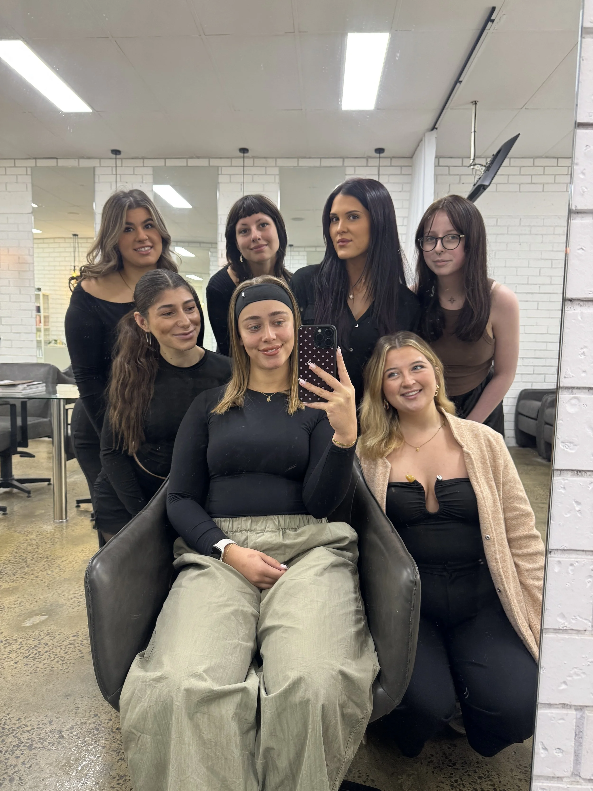 Group of seven women taking a selfie in front of a mirror in a modern indoor setting with white brick walls and ceiling lights.