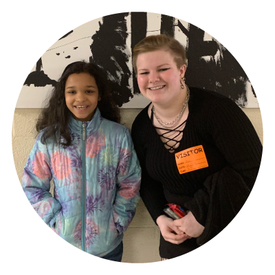 Two smiling girls standing in front of a black and white abstract painting. The girl on the left has long, dark curly hair and is wearing a colorful patterned jacket. The girl on the right has blonde hair tied back and is wearing a black top and an orange visitor badge.
