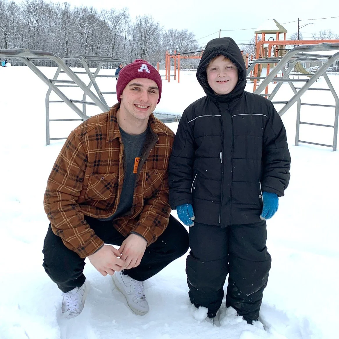 A young man in a brown plaid jacket and maroon beanie kneels next to a young boy in a black winter coat and blue gloves in a snowy park, with playground equipment in the background.