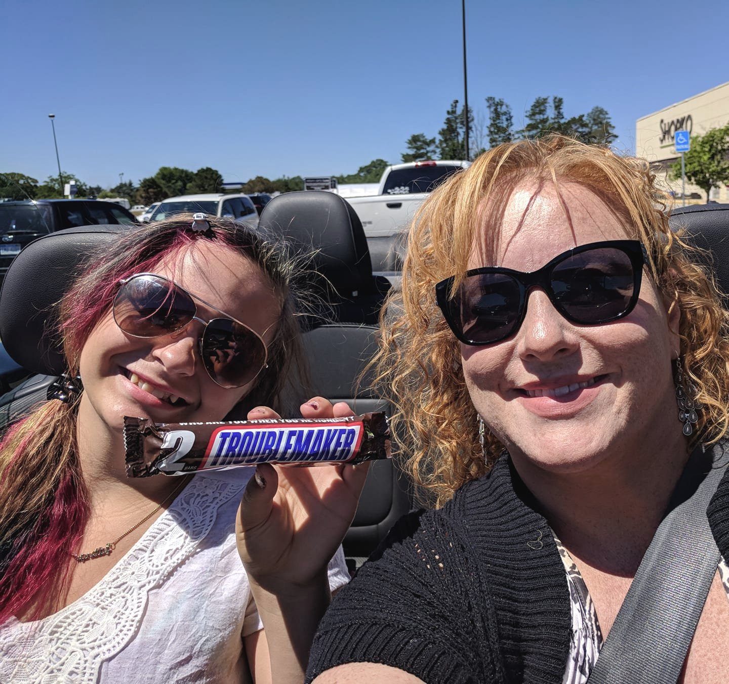 Two women smiling in a car, one holding a Trouble Maker energy bar, both wearing sunglasses, with cars in the parking lot and a shopping center in the background.