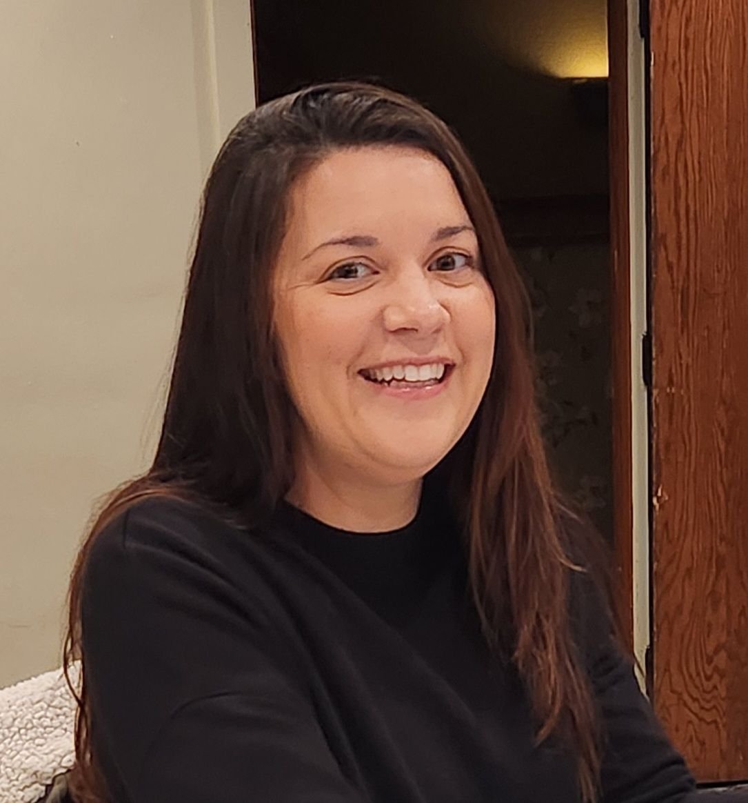 A woman with long brown hair smiling, wearing a black top, indoors.