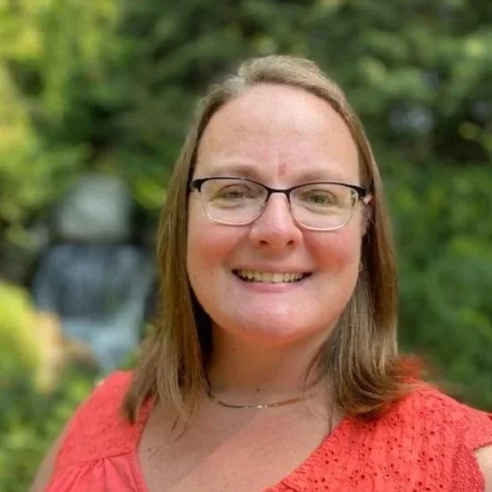 A woman wearing glasses and a red top outdoors, smiling with greenery and a blurred waterfall in the background.