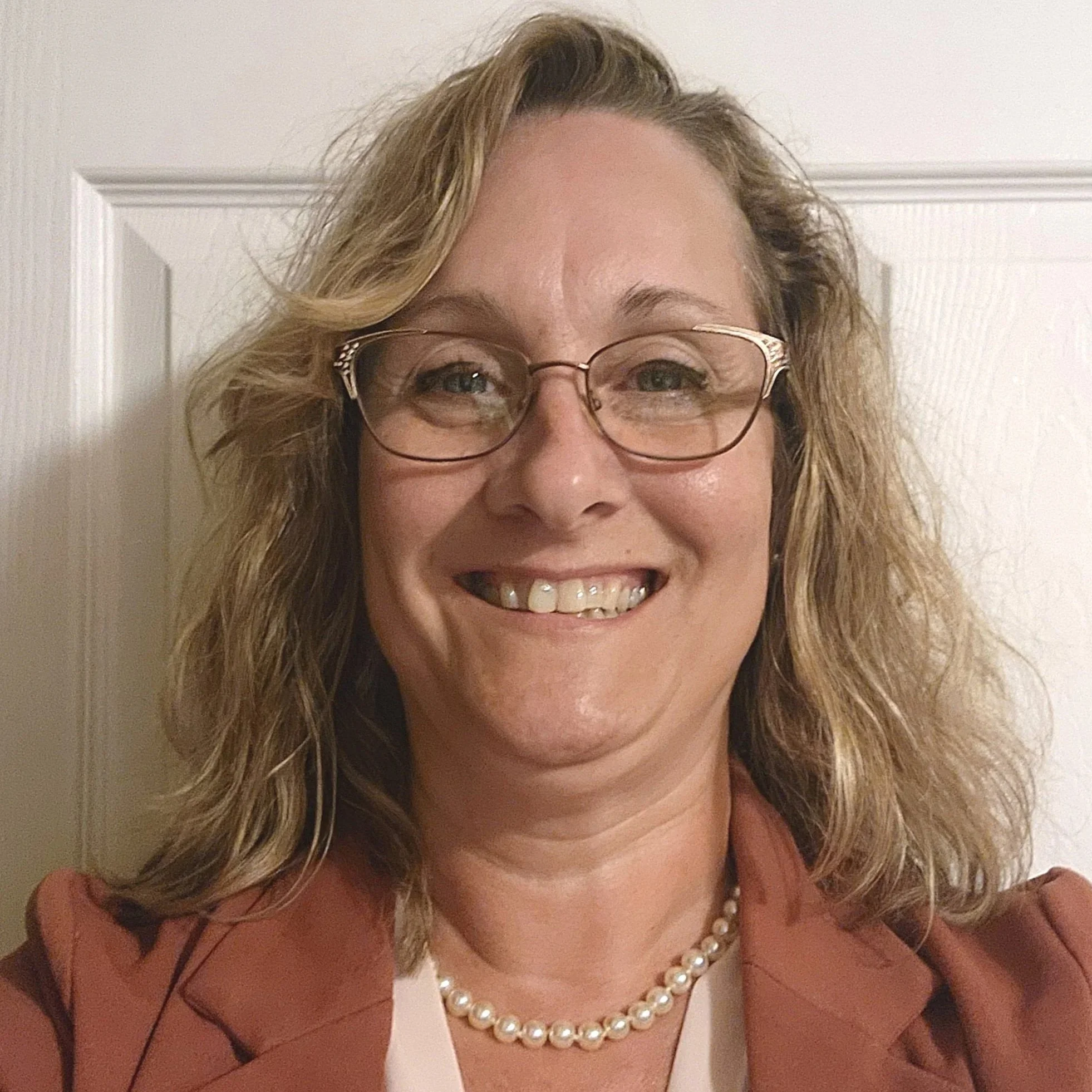 Close-up of a woman with blonde, wavy hair, glasses, wearing a pearl necklace, smiling, and dressed in a brown top with black accents.