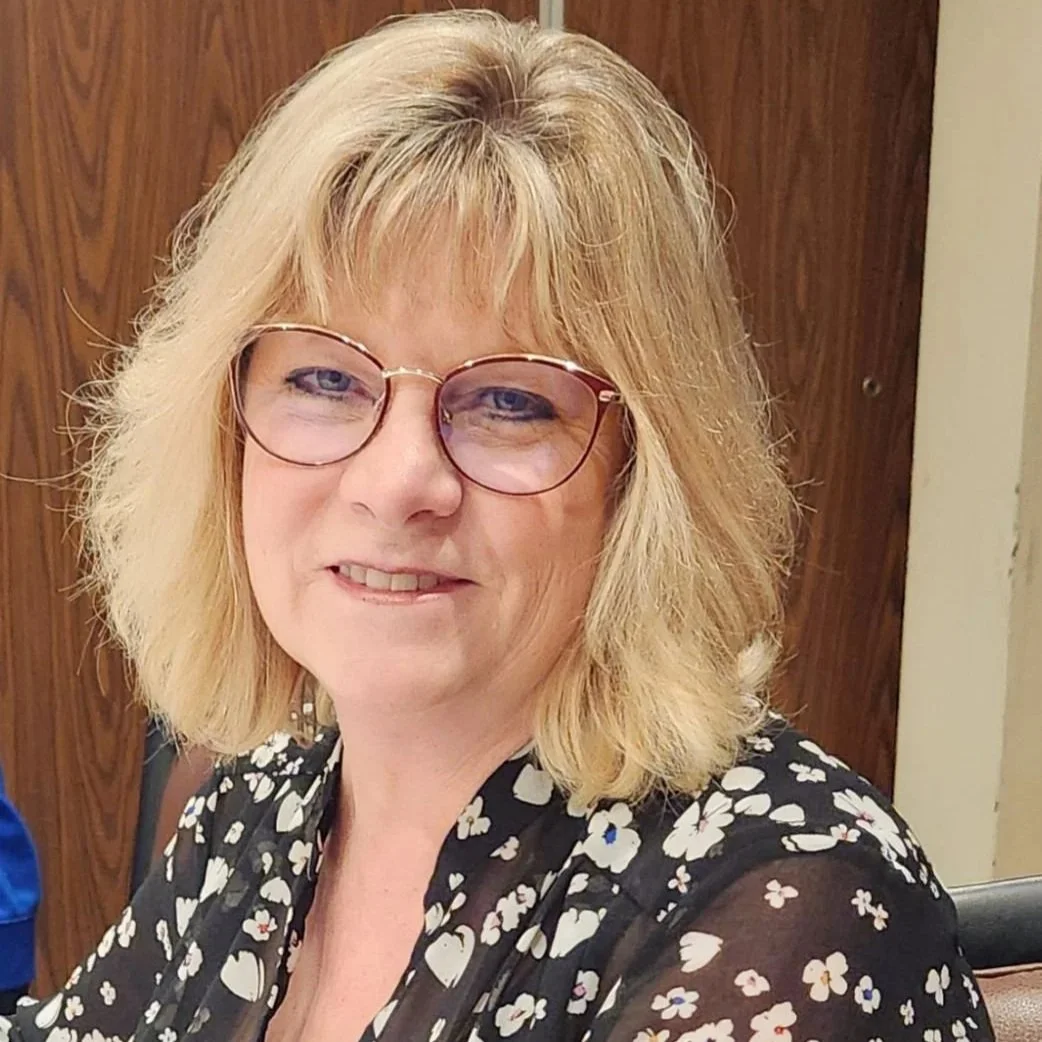 A middle-aged woman with blonde, shoulder-length hair wearing glasses and a black blouse with a white floral pattern, smiling at the camera.