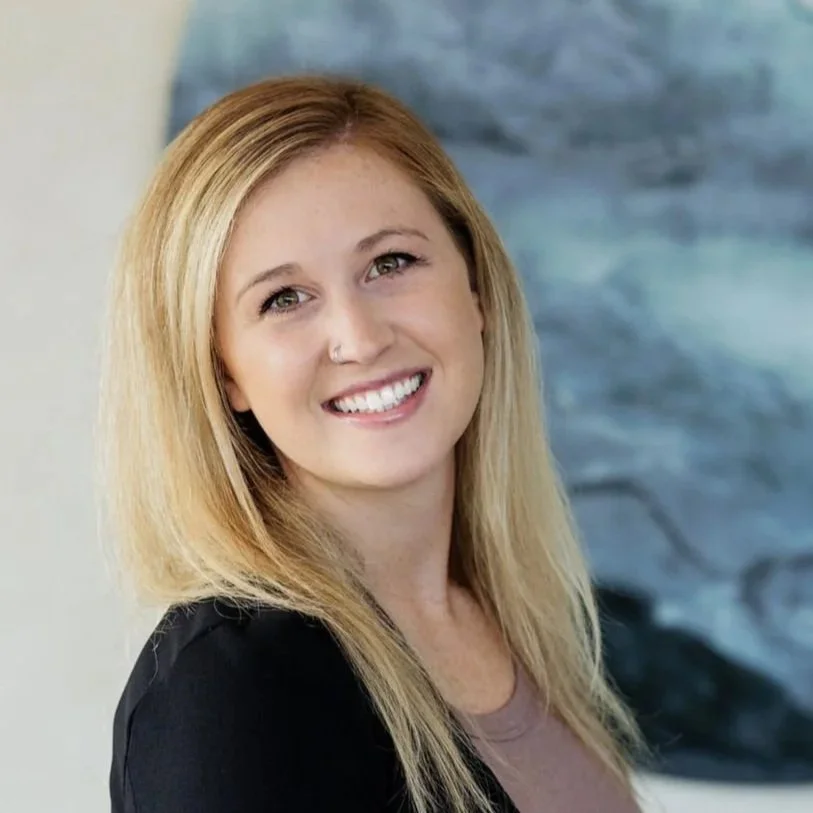 A woman with blonde hair, smiling, wearing a black blazer and a beige top, standing in front of a blurry blue and green background.