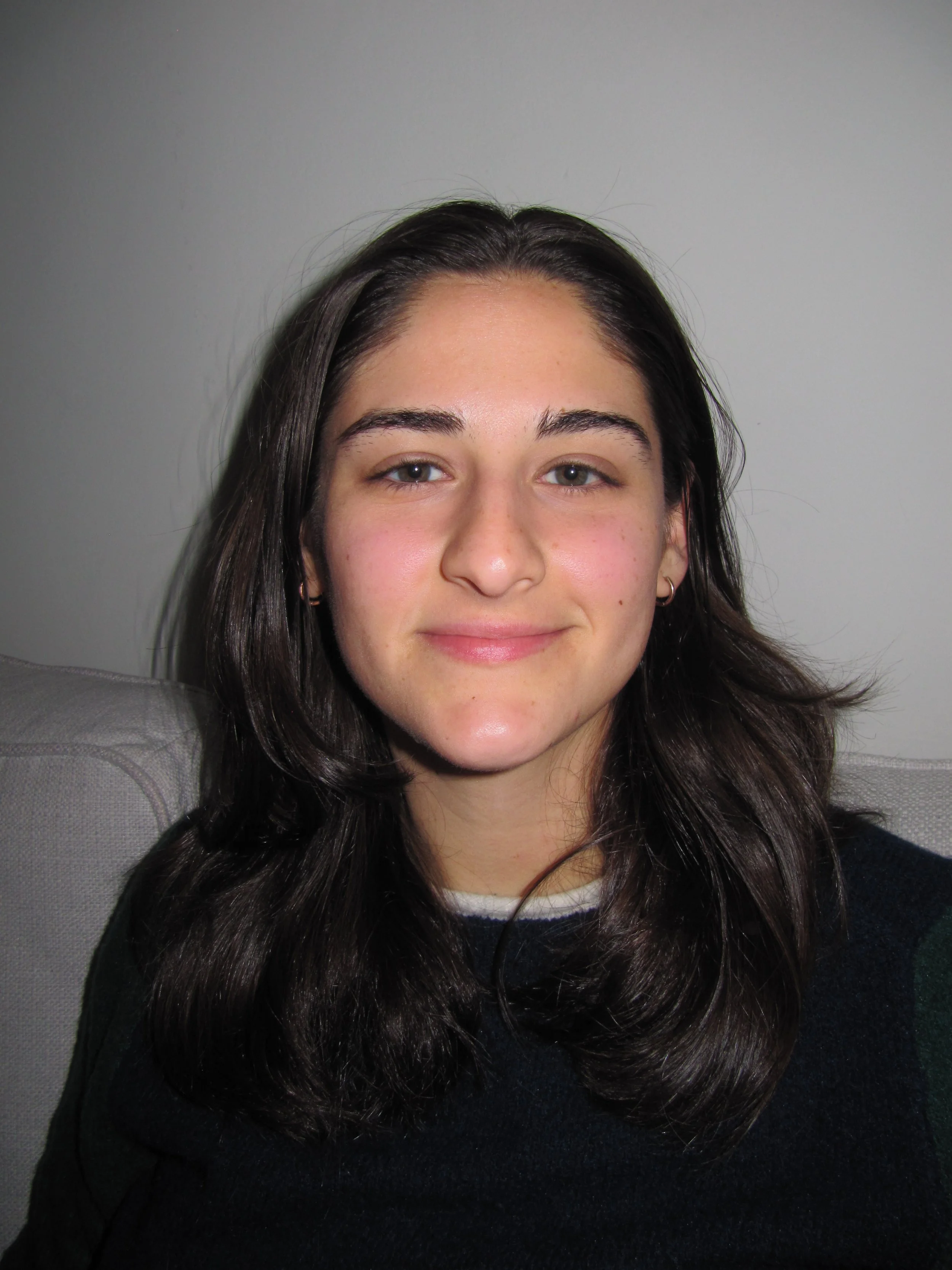 A young woman with long dark hair, light skin, and wearing a dark sweater, smiling slightly, sitting on a gray couch against a plain white wall.
