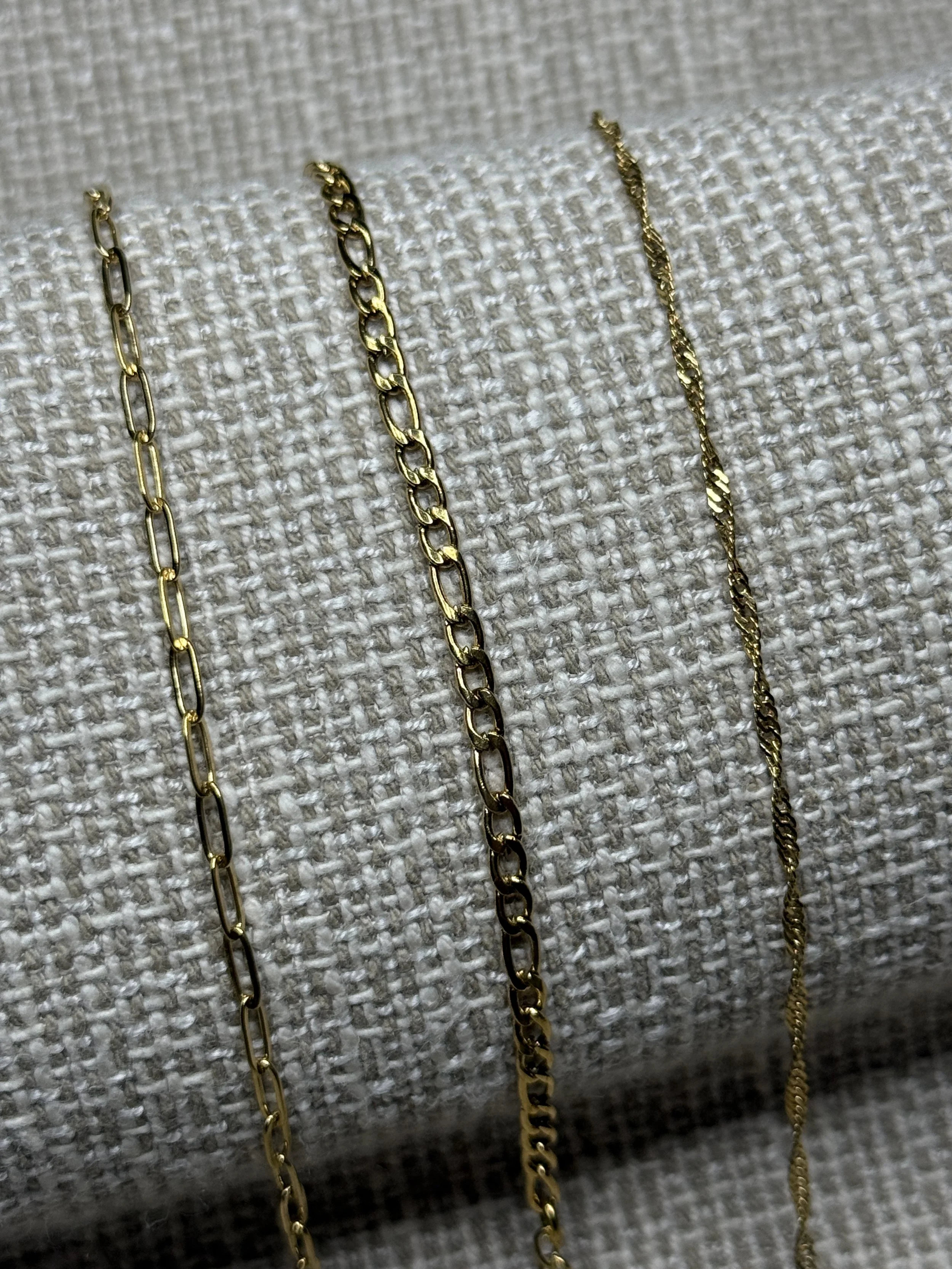Close-up of three delicate gold chain necklaces laid out on a textured fabric background.