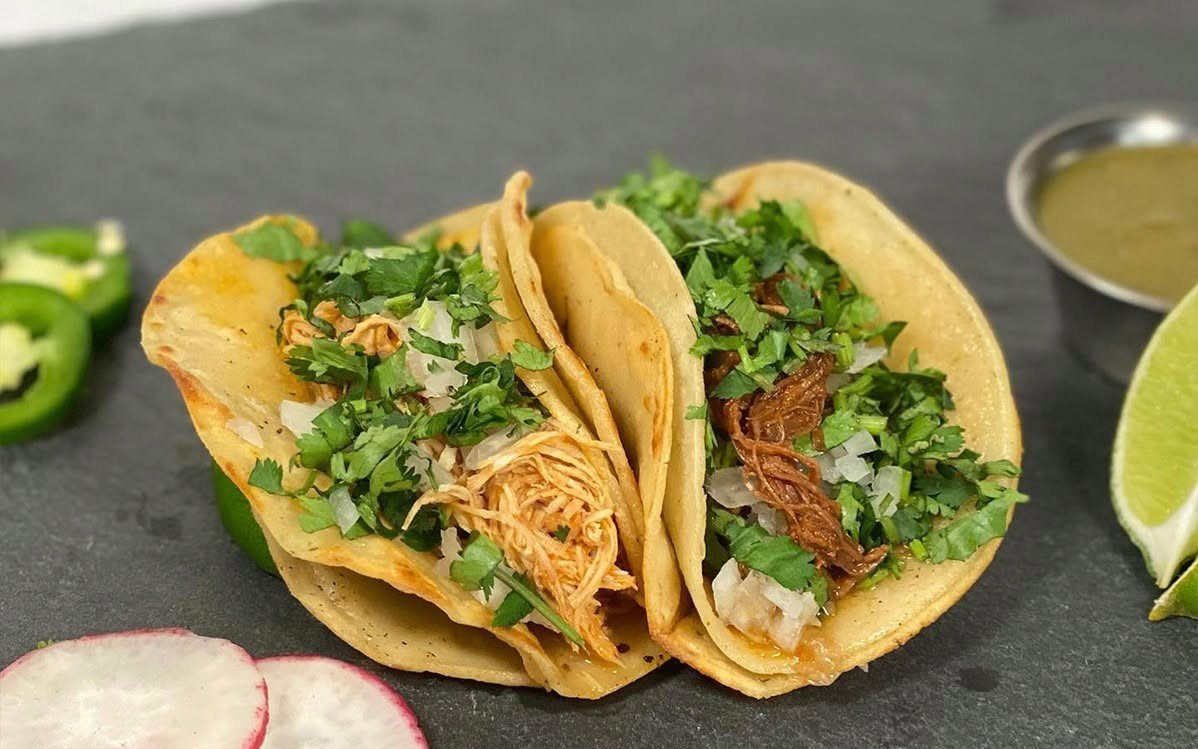 Two soft taco shells filled with shredded chicken, chopped cilantro, diced onions, and shredded beef on a dark surface with lime wedges, radish slices, jalapeño slices, and a small cup of green sauce.