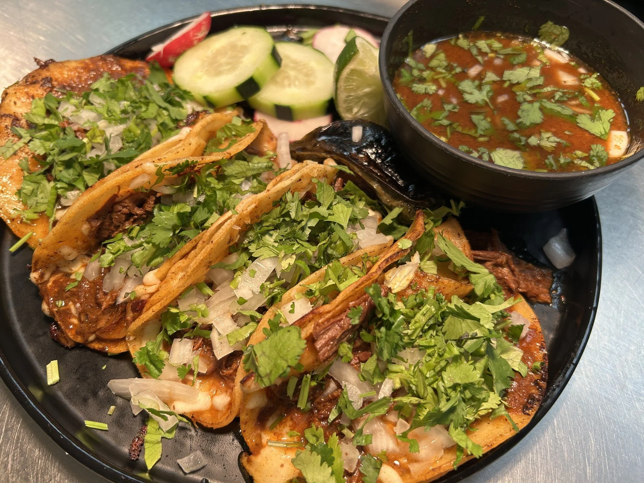 Plate of grilled fish tacos topped with chopped cilantro, with sliced cucumbers, radishes, and lime wedges on the side, and a small bowl of salsa garnished with cilantro.