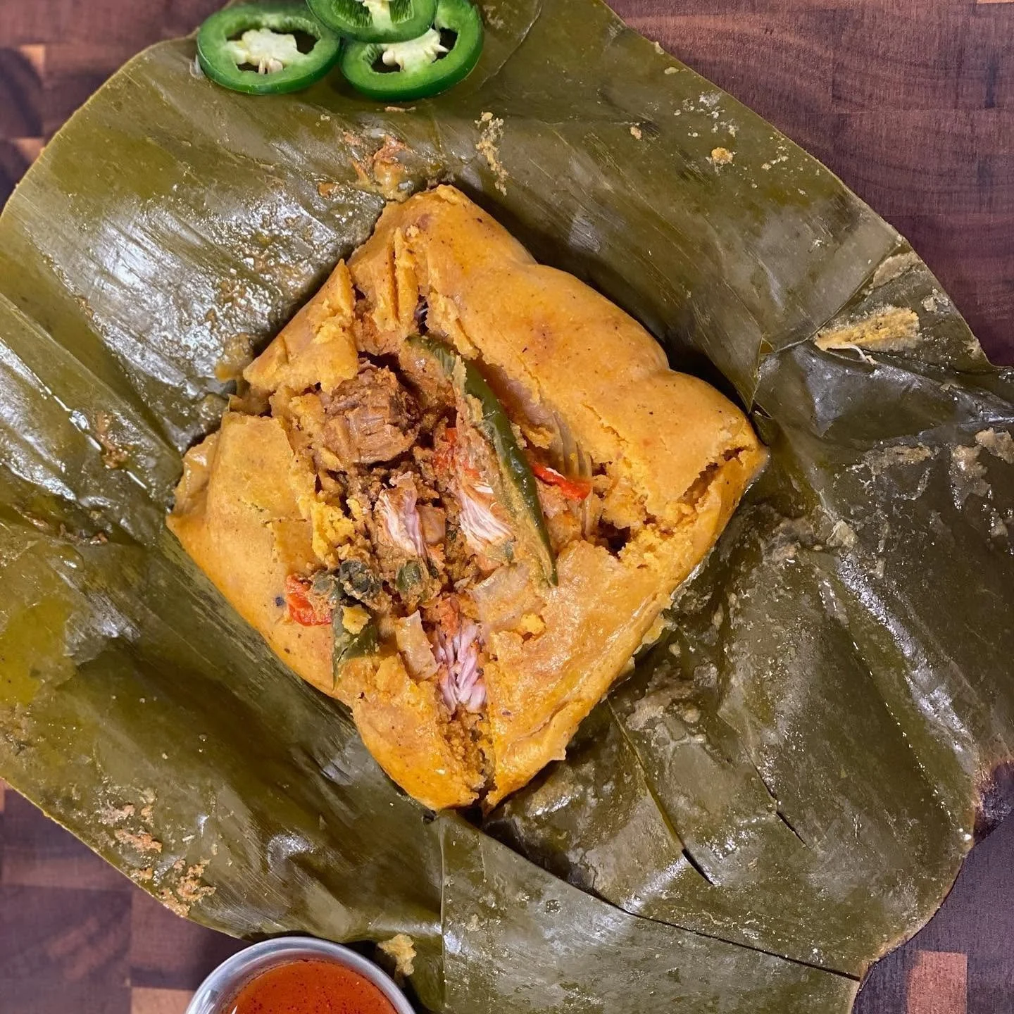 A traditional Mexican tamale wrapped in corn husk, partially opened to show filling of shredded meat, vegetables, and spices. Sliced jalapenos are on top, with a small cup of red sauce at the bottom.